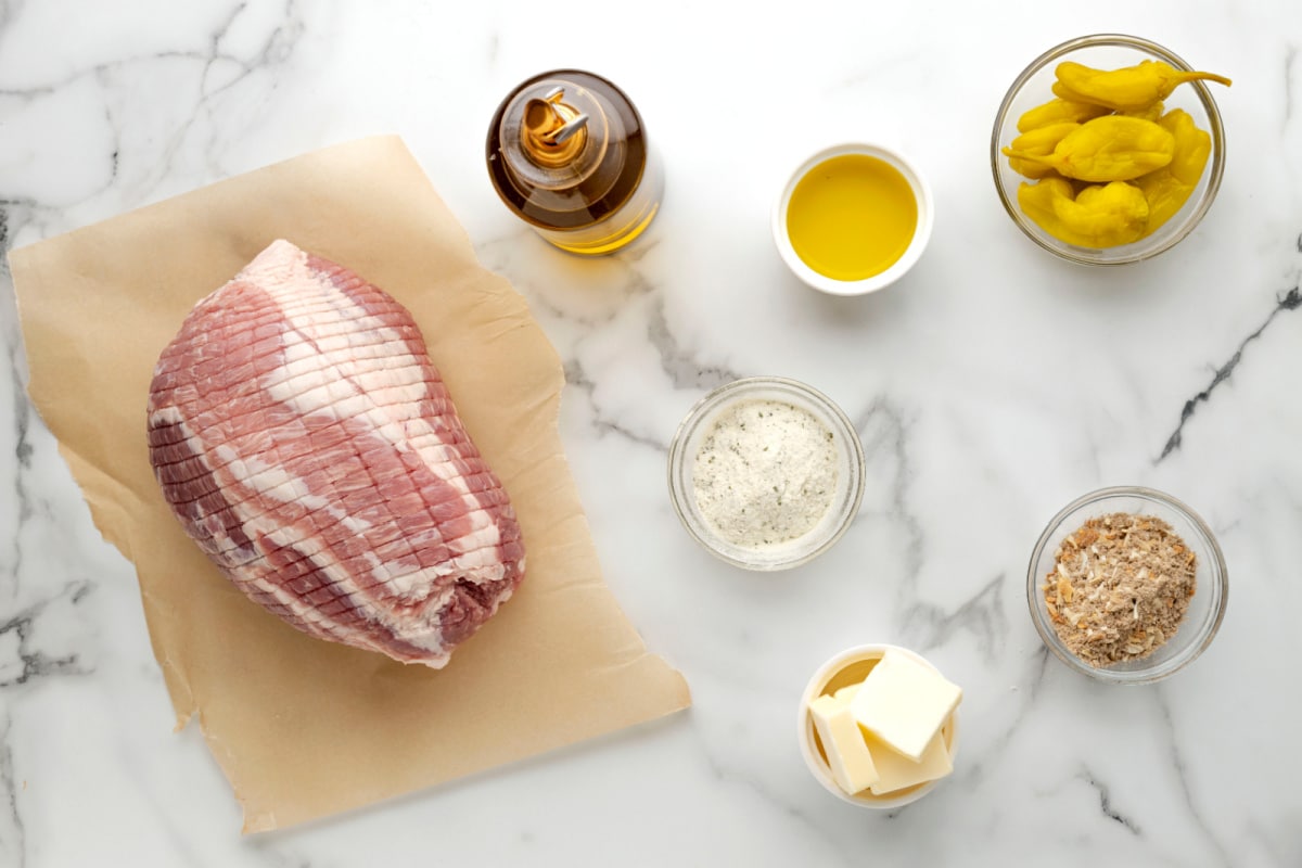 Ingredients for slow cooker Mississippi pork roast including raw pork loin, ranch seasoning, onion soup mix, butter, olive oil, and pepperoncini.