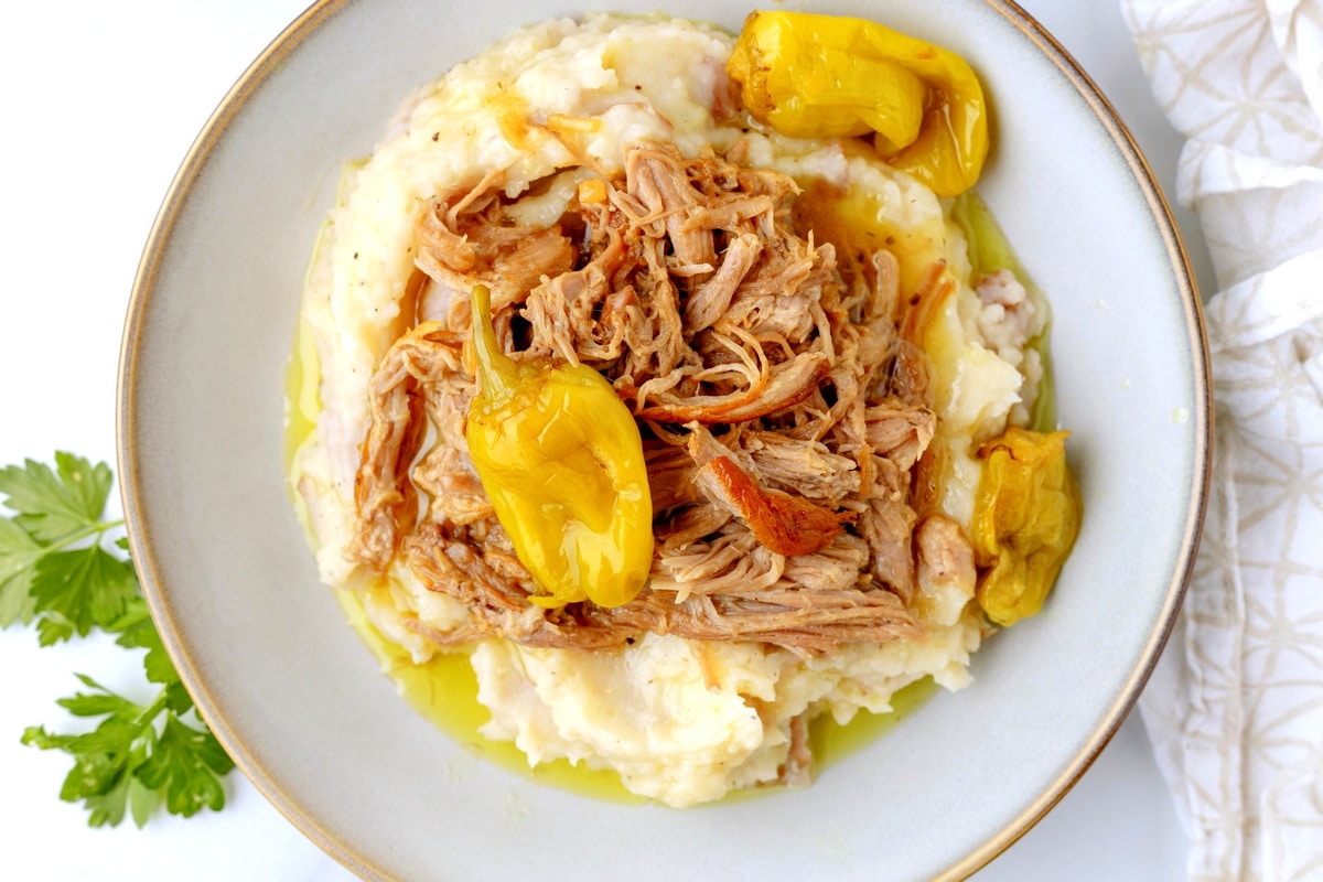 Overhead view of shredded Mississippi pork loin served on mashed potatoes with yellow pepperoncini peppers and cooking liquid.