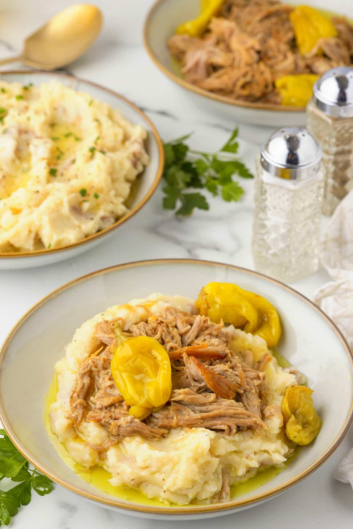 Slow cooker Mississippi pork roast on mashed potatoes with pepperoncini peppers, with additional mashed potatoes and pork in the background.