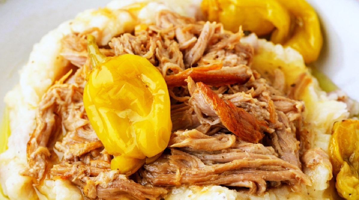 Close-up of shredded Mississippi pork roast with browned strands of pork, whole pepperoncini peppers, and mashed potatoes.