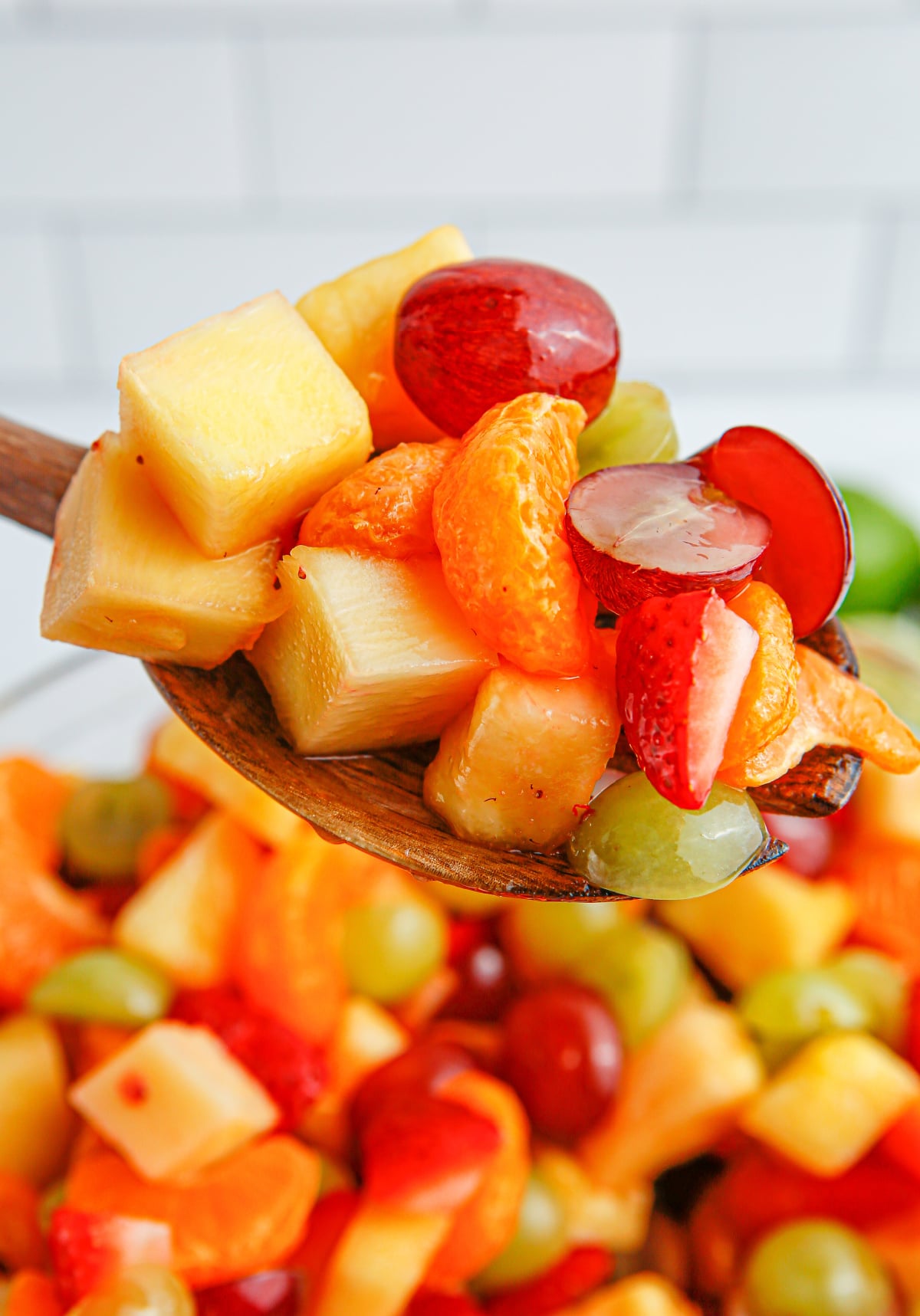 Honey Lime Fruit Salad scooped by a wooden spoon Close-up of a wooden spoon scooping up a portion of the honey lime fruit salad, showing a mix of colorful, glossy fruit pieces.