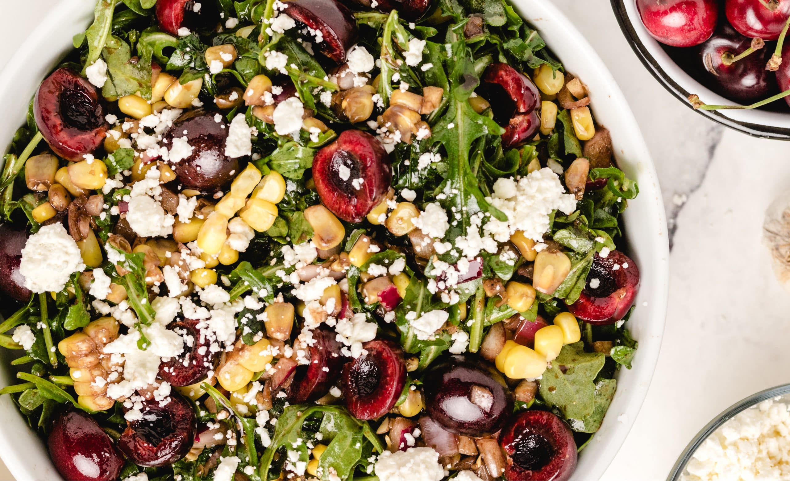 cherry corn salad with arugula and feta Overhead view of cherry corn salad with arugula, feta, and fresh sweet cherries in a white bowl.