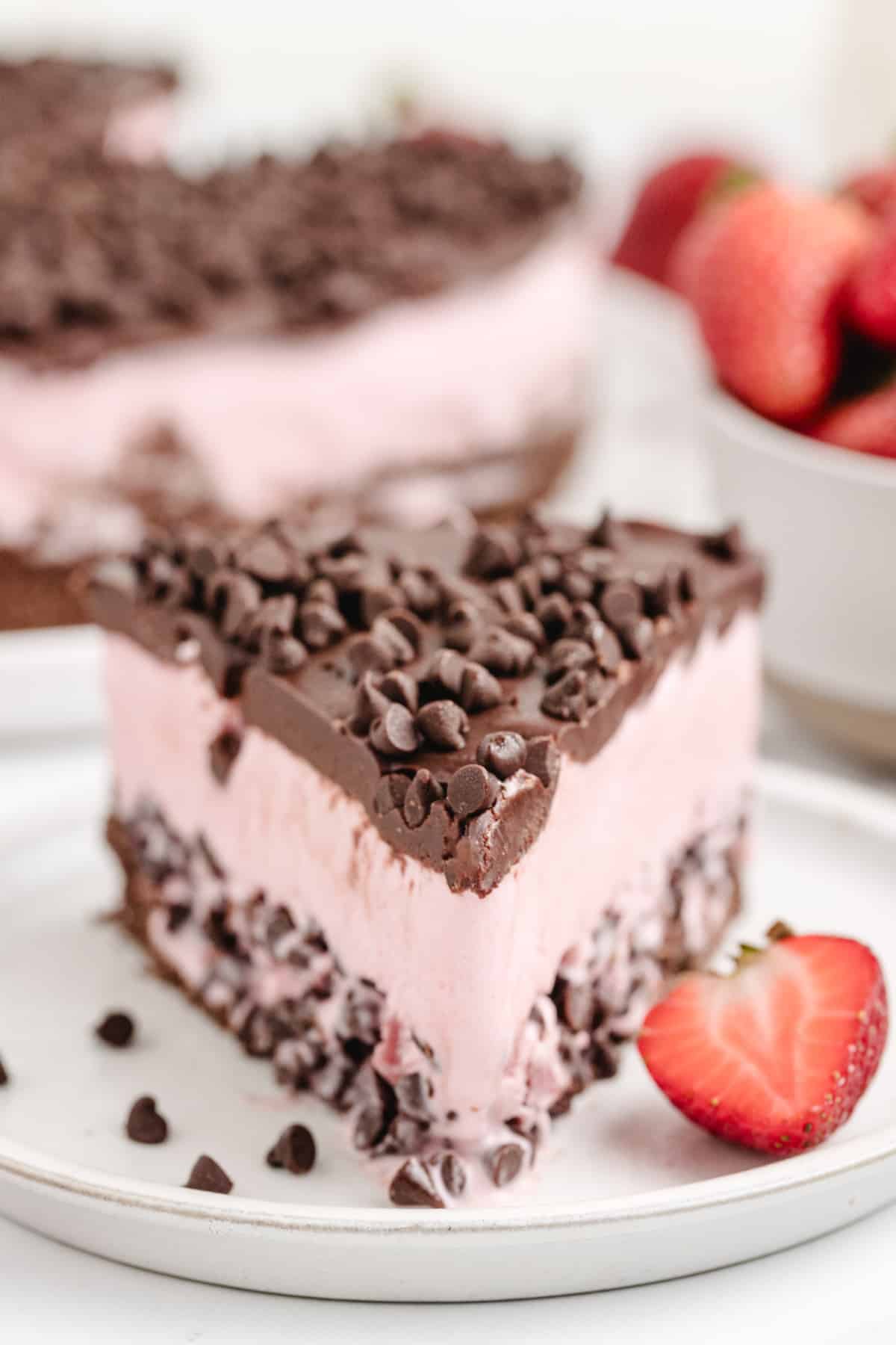 a piece of Strawberry Chocolate Chip Ice Cream Cake A creamy and cold slice of strawberry chocolate chip ice cream cake on a plate, showcasing its layers of chocolate graham cracker crust, strawberry ice cream with chocolate chips, and rich chocolate topping.