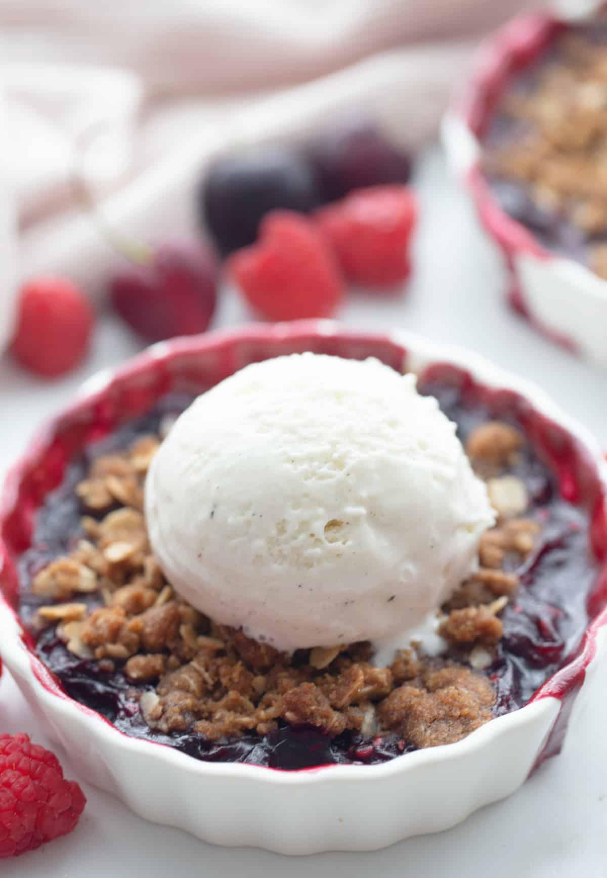 Fresh cherry berry crisp topped with vanilla ice cream in a white ramekin, with raspberries and cherries in the background.