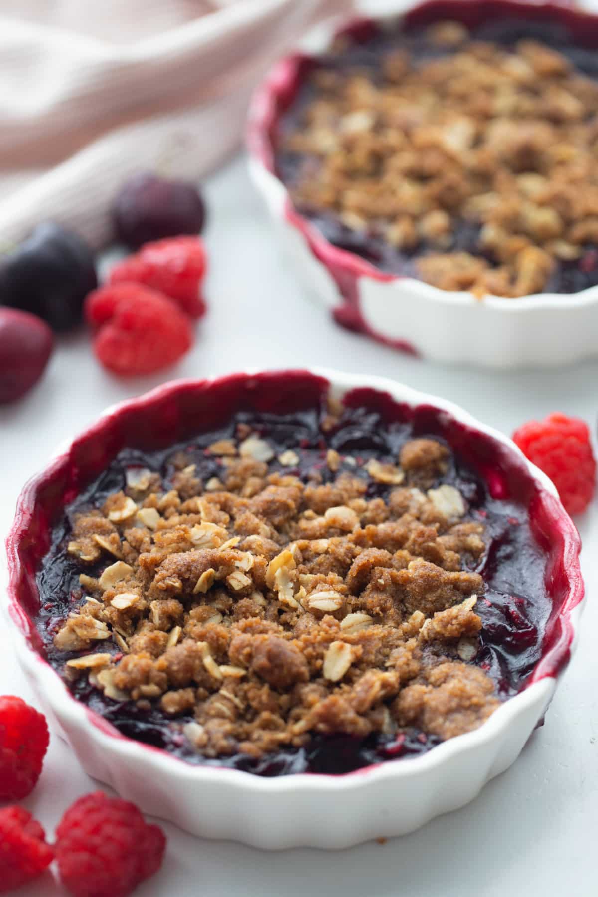 Two servings of fresh cherry crisp with oat topping in white dishes, surrounded by fresh raspberries and cherries.