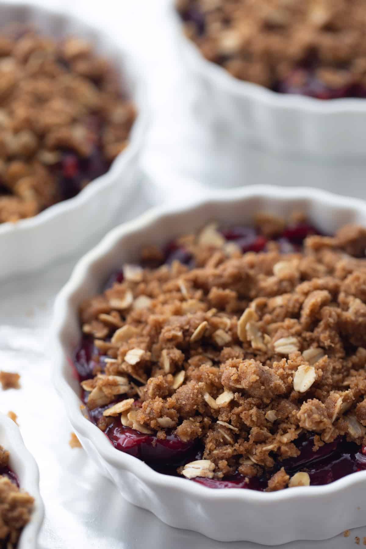 Fresh cherry crisp with oat topping layered over fruit filling in white ramekins on foil-lined tray. 