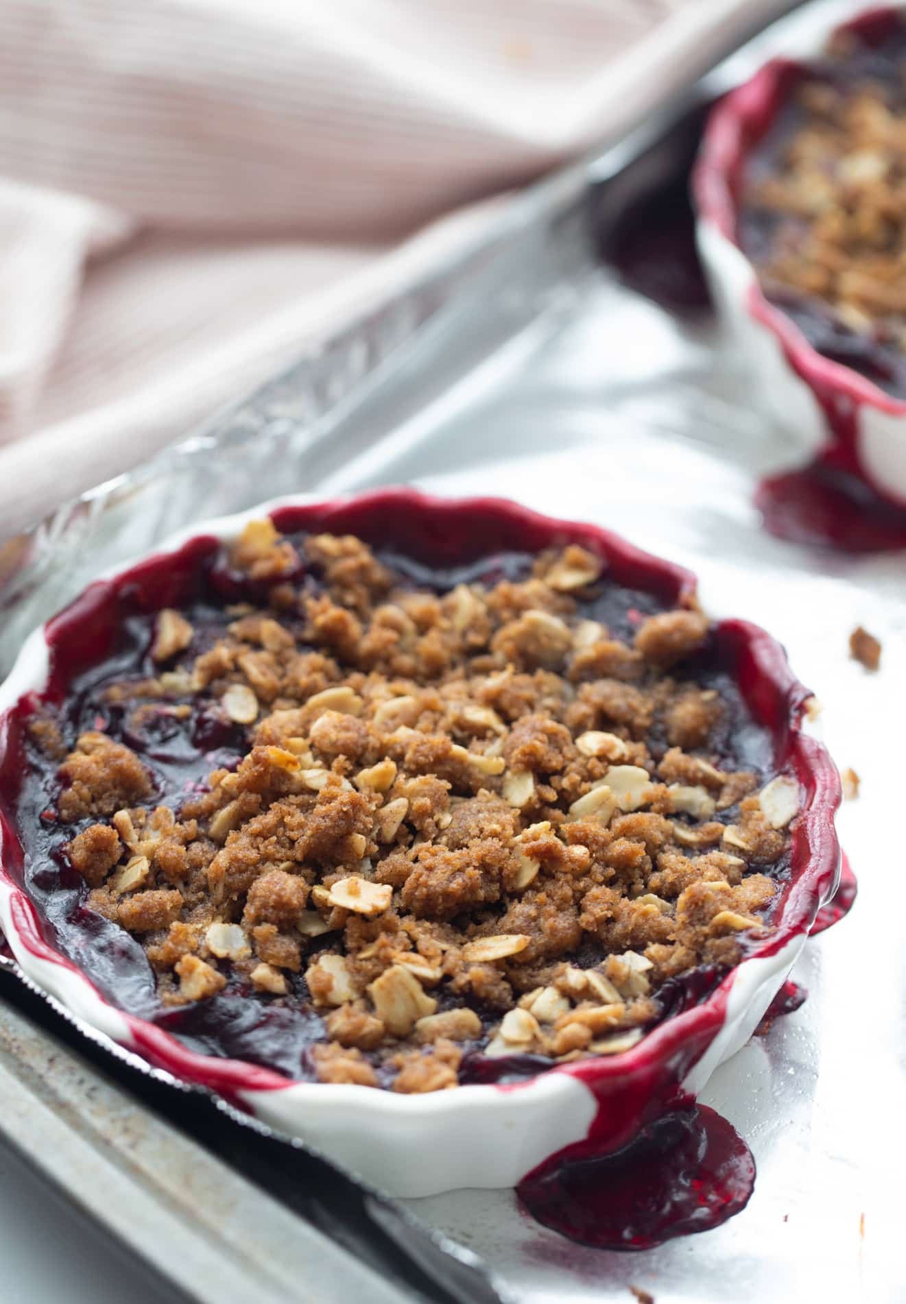 Baked fresh cherry crisp with oat topping in ramekin, fruit bubbling over onto foil-lined tray.