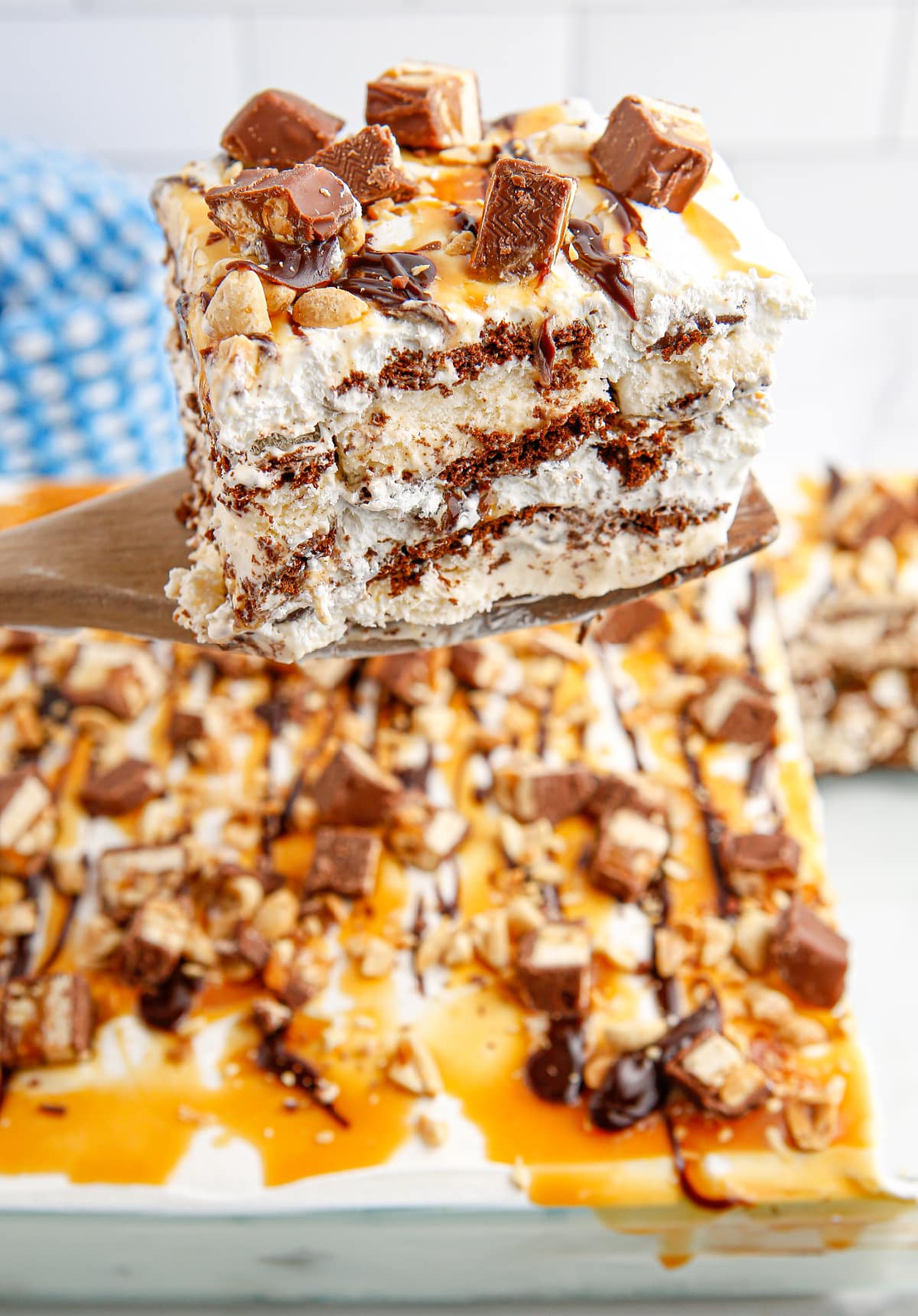 snickers ice cream cake slice on spatula with full dessert in background Large square of snickers ice cream cake on spatula above a full pan topped with candy and sauces