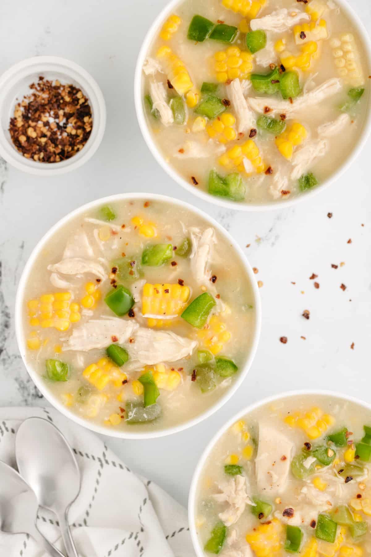 Three bowls of chicken corn chowder topped with shredded chicken, fresh sweet corn, green bell peppers, and crushed red pepper flakes.