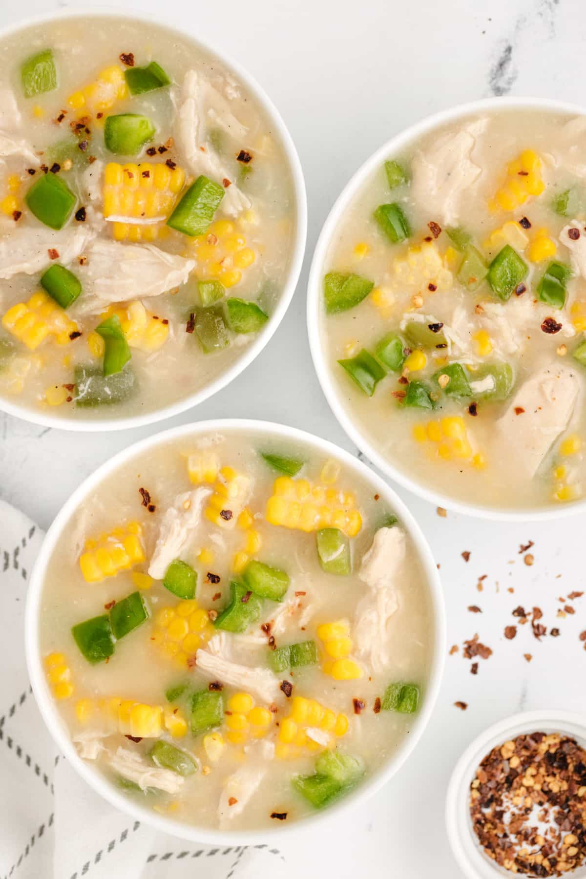 Chicken corn chowder with shredded chicken, fresh sweet corn, green bell peppers, and crushed red pepper flakes served in three bowls.