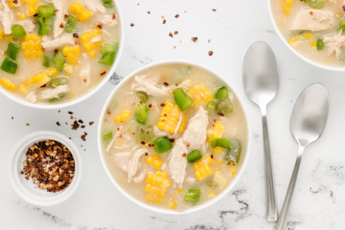 Chicken corn chowder served in individual bowls with shredded chicken, fresh sweet corn, green bell peppers, and crushed red pepper flakes.