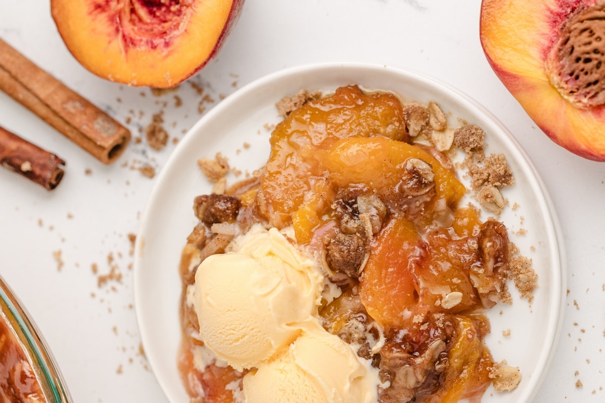 Bowl of warm peach crisp topped with vanilla ice cream, surrounded by fresh peaches and cinnamon sticks.