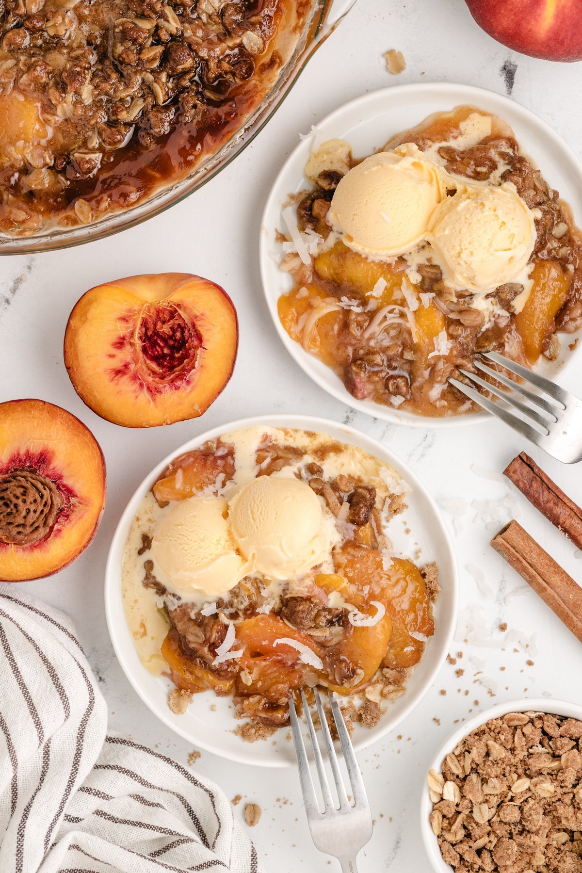 Two plates of peach crisp with ice cream, fresh peaches, and cinnamon sticks on table.