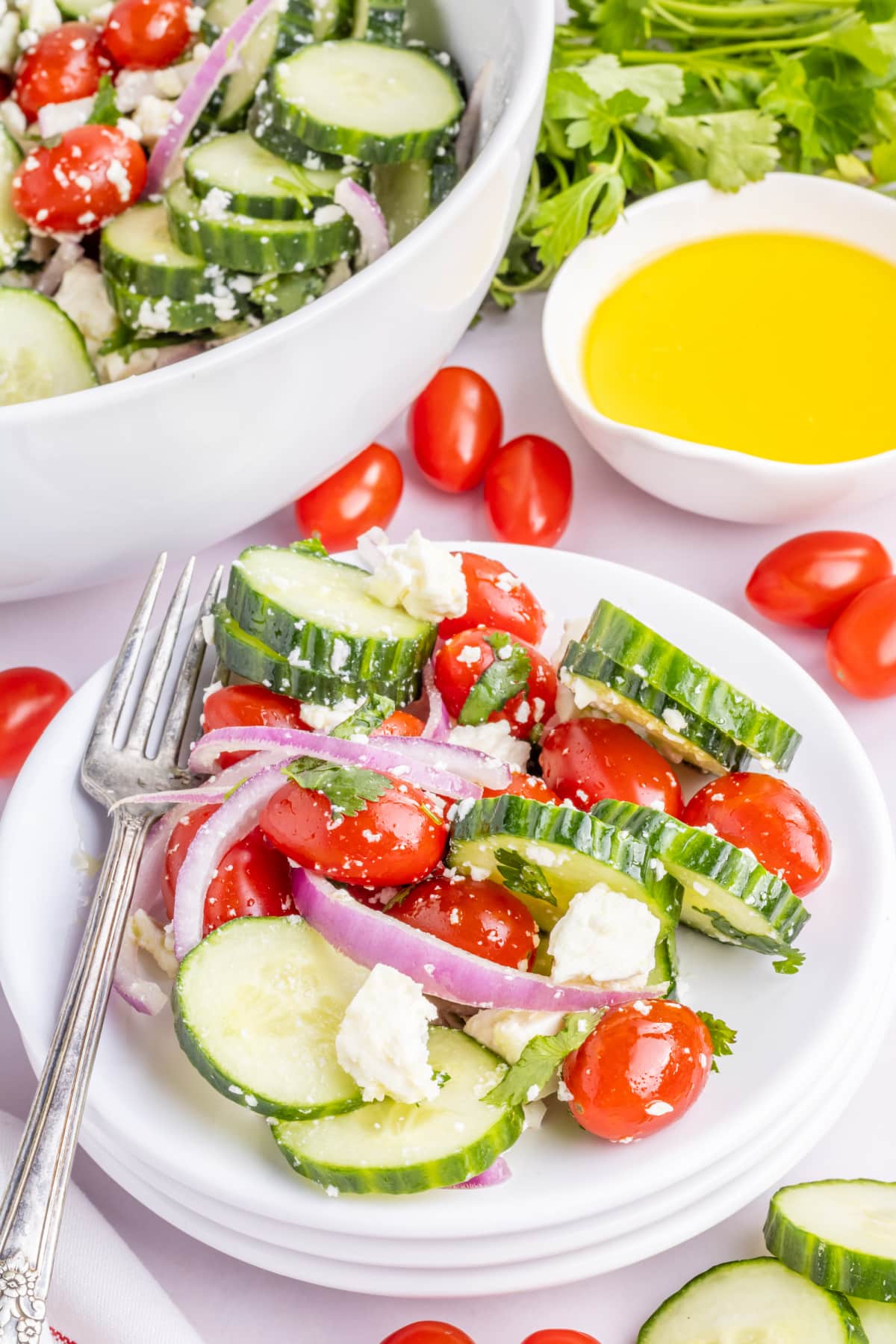 Cold Cucumber Salad with Feta and Lemon Dressing Side view of a small plate of cucumber feta salad with tomatoes and red onion, with parsley and olive oil nearby.