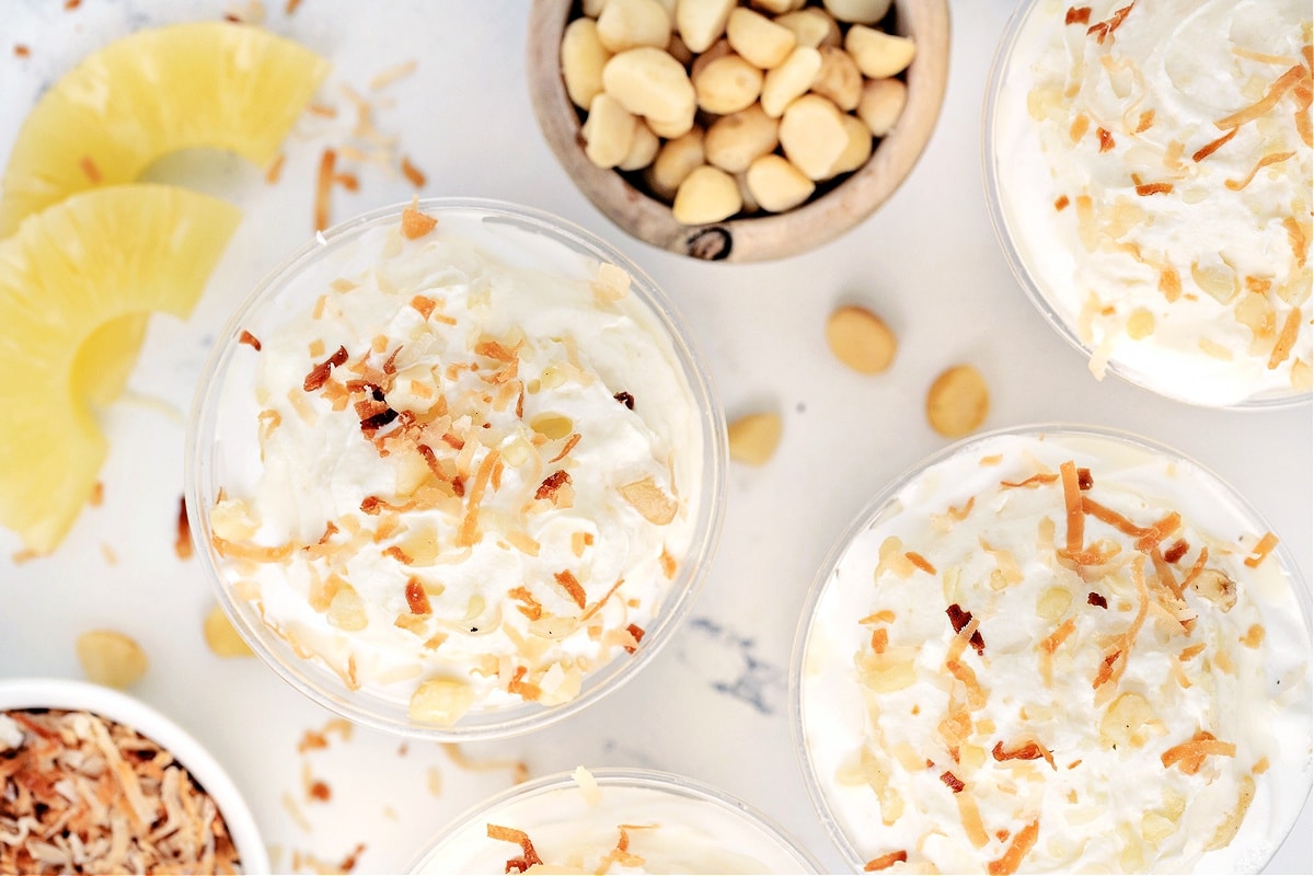 Overhead view of Hawaiian Dream Dessert Cups with toasted coconut and macadamias Overhead view of Hawaiian Dream Dessert Cups topped with whipped cream, toasted coconut, and macadamia nuts surrounded by pineapple slices.