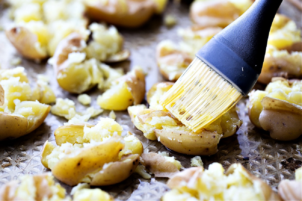 A pastry brush coating hot smashed potatoes with apple cider vinegar on a sheet pan, the final step that gives salt and vinegar smashed potatoes their tang.
