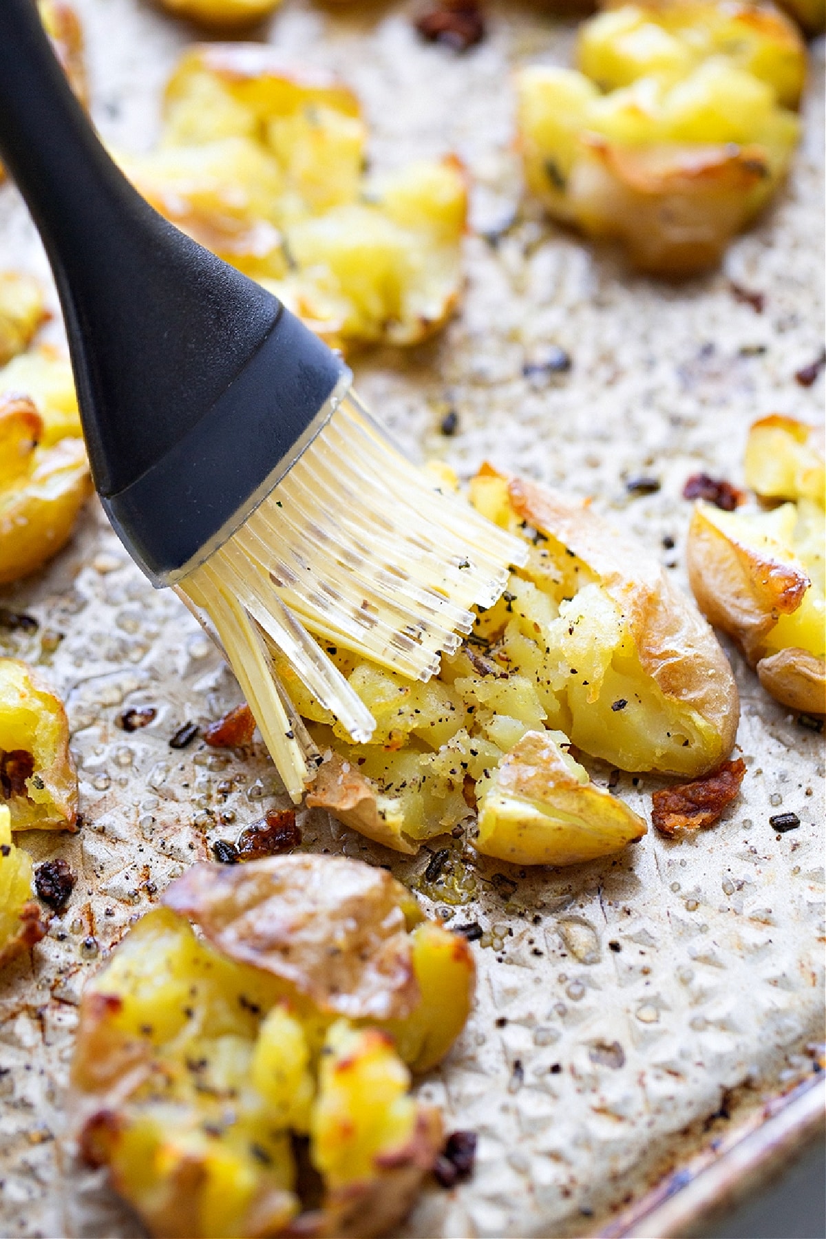 A pastry brush adding apple cider vinegar to hot smashed potatoes on a sheet pan, giving salt and vinegar smashed potatoes their tang.