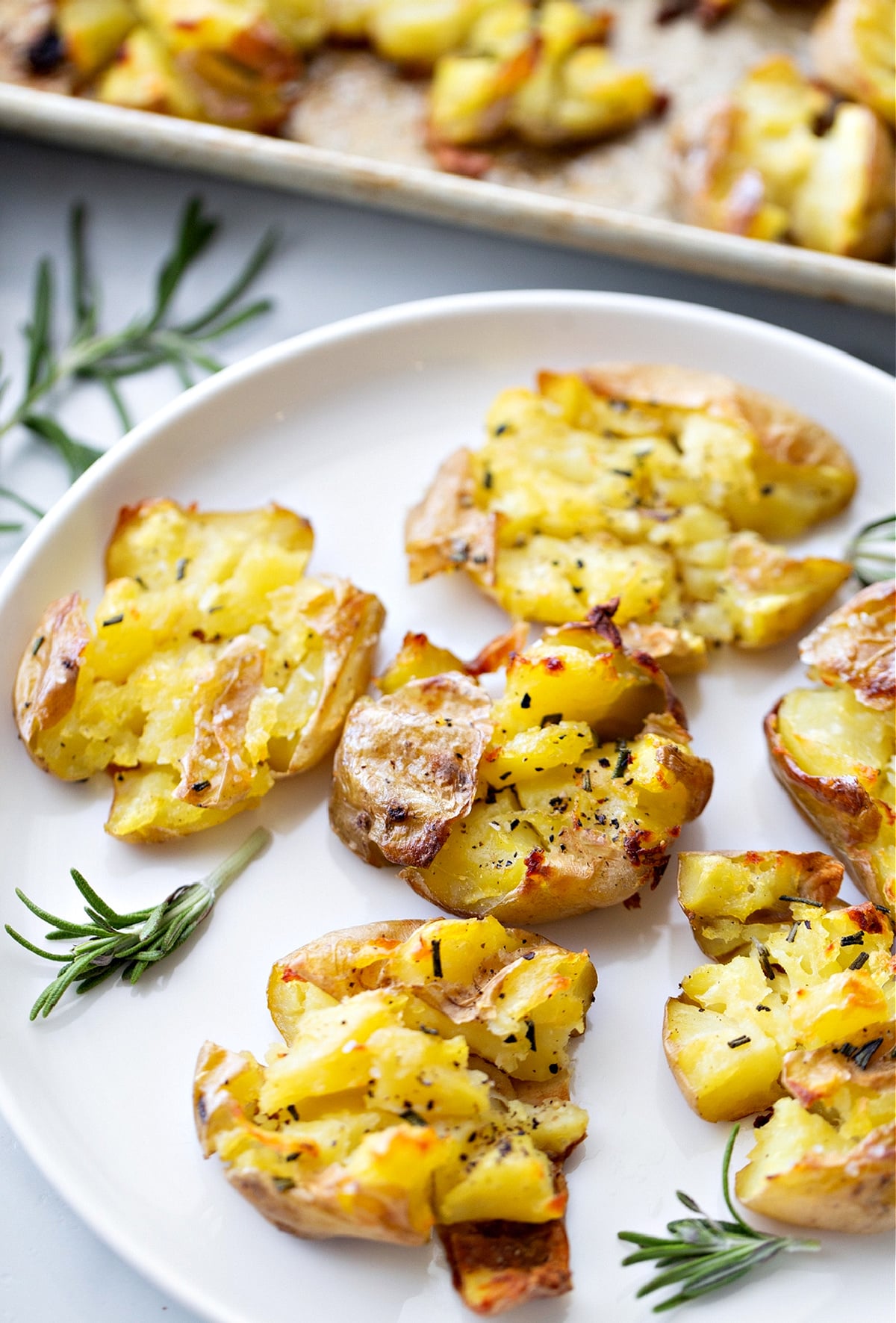 Salt and vinegar smashed potatoes with crisp edges and rosemary on a white plate, showing their roasted texture, by Noble Pig.