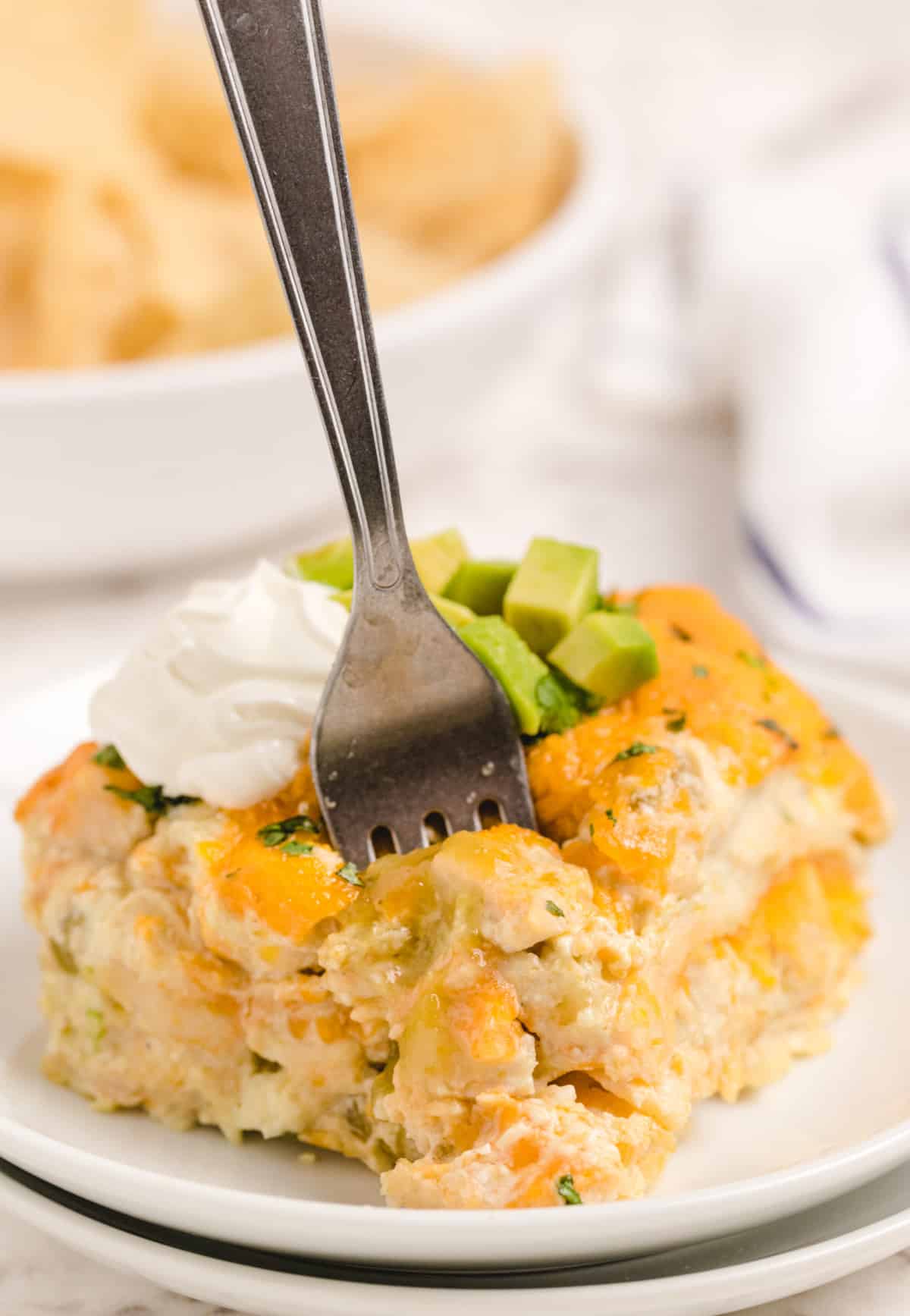 Fork cutting into a slice of Green Chile Chicken Enchilada Casserole Fork spearing the top of a slice of Green Chile Chicken Enchilada Casserole with avocado and sour cream