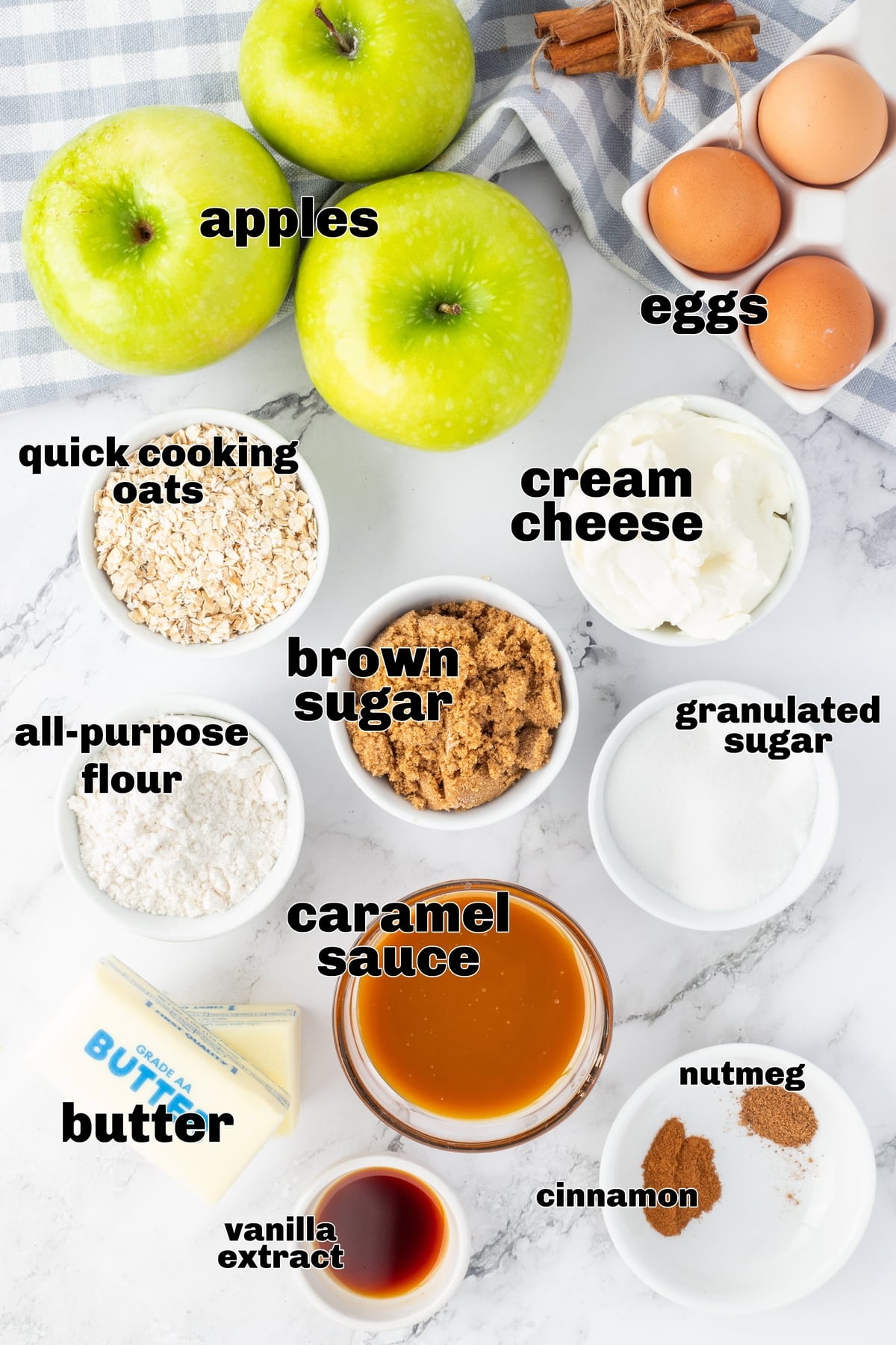 Apple cheesecake bars ingredients. Ingredients for apple cheesecake bars laid out on a marble counter, including apples, cream cheese, butter, oats, sugar, and caramel sauce.