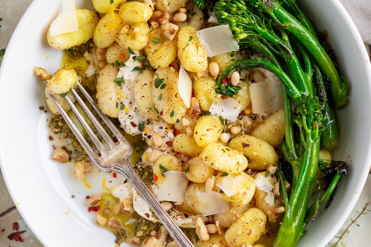 Close-up of crispy gnocchi with burrata, broccolini, toasted nuts, and pesto in a white bowl with a fork.