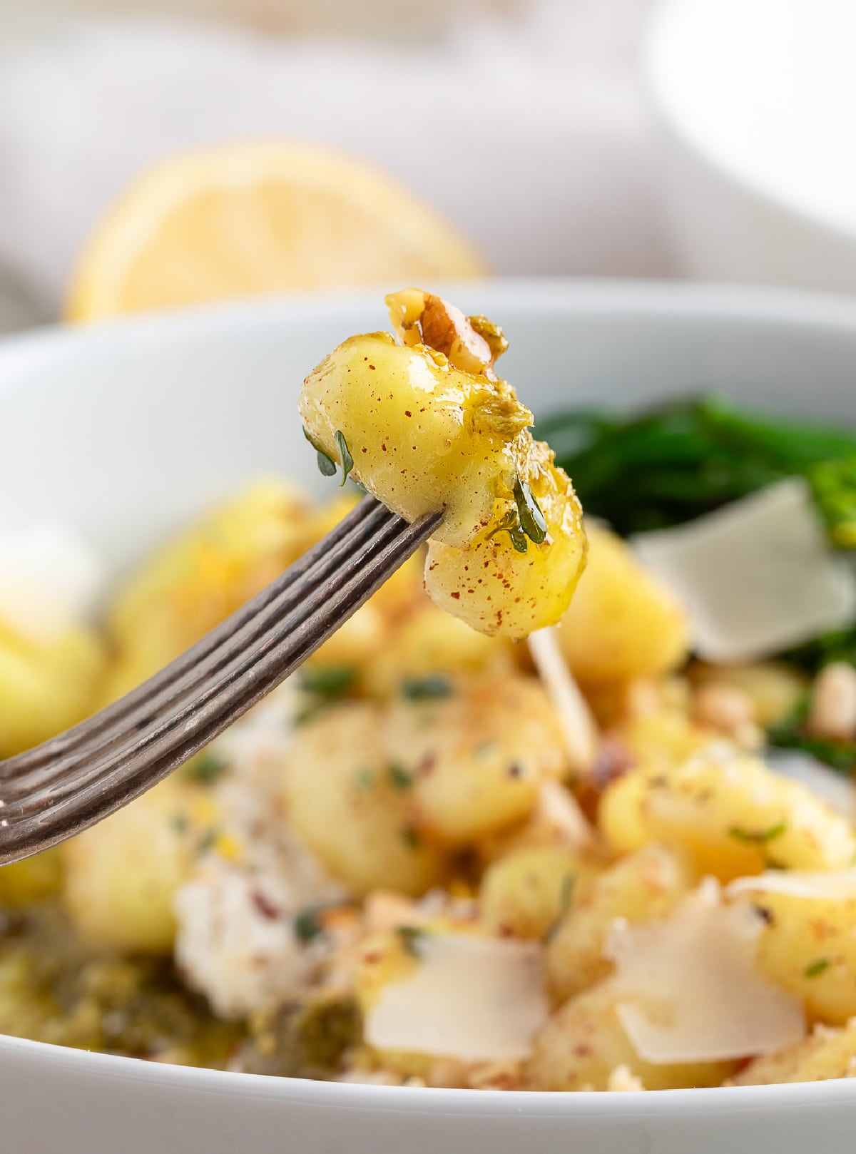 Fork holding a bite of gnocchi coated in brown butter with fresh herbs, over a bowl with burrata, pesto, shaved parmesan, and broccolini.