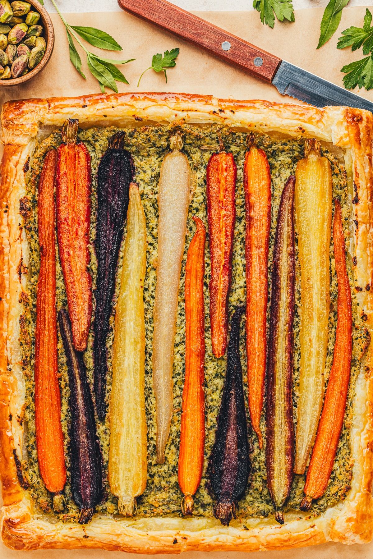 A fully baked carrot tart with pistachio pesto and ricotta on a parchment-lined baking sheet, showing its golden puff pastry crust.