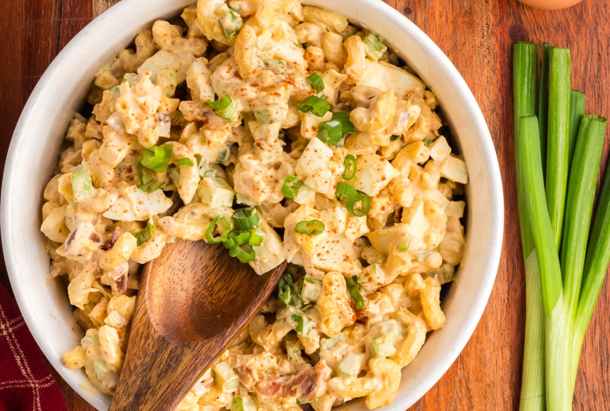Deviled Egg Pasta Salad with green onion. A bowl of deviled egg pasta salad with a wooden spoon resting on the side and fresh green onions beside it.