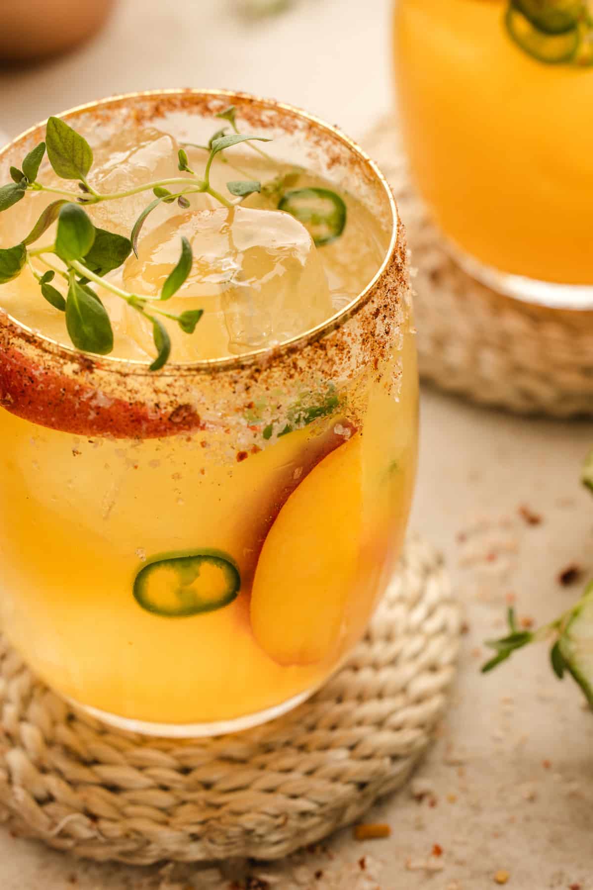 Close-up of the Hot Honey Peach Margarita in a glass with a chili salt rim, garnished with peach slices, jalapeño, and a thyme sprig.