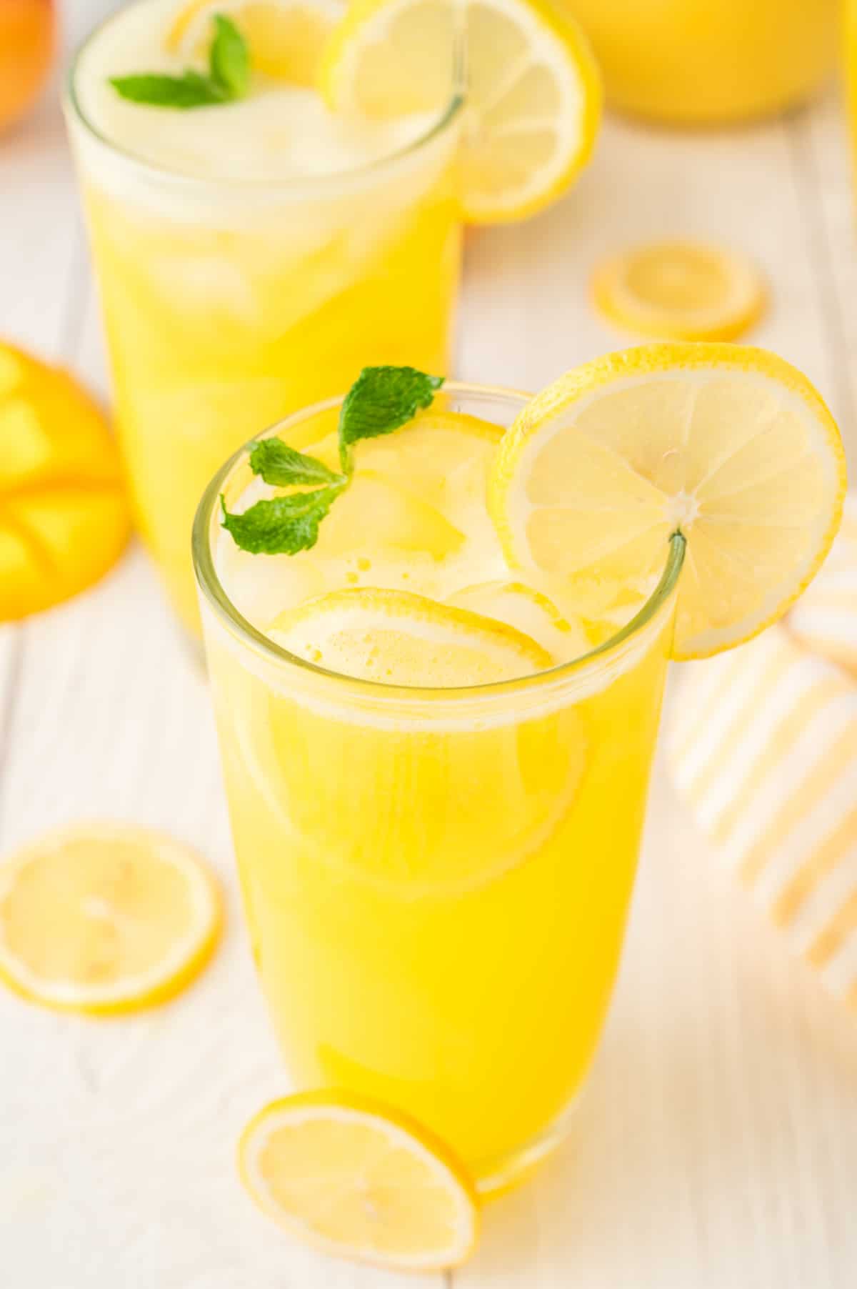 Two glasses of mango lemonade on a table, garnished with lemon slices and mint, with condensation forming on the glasses.