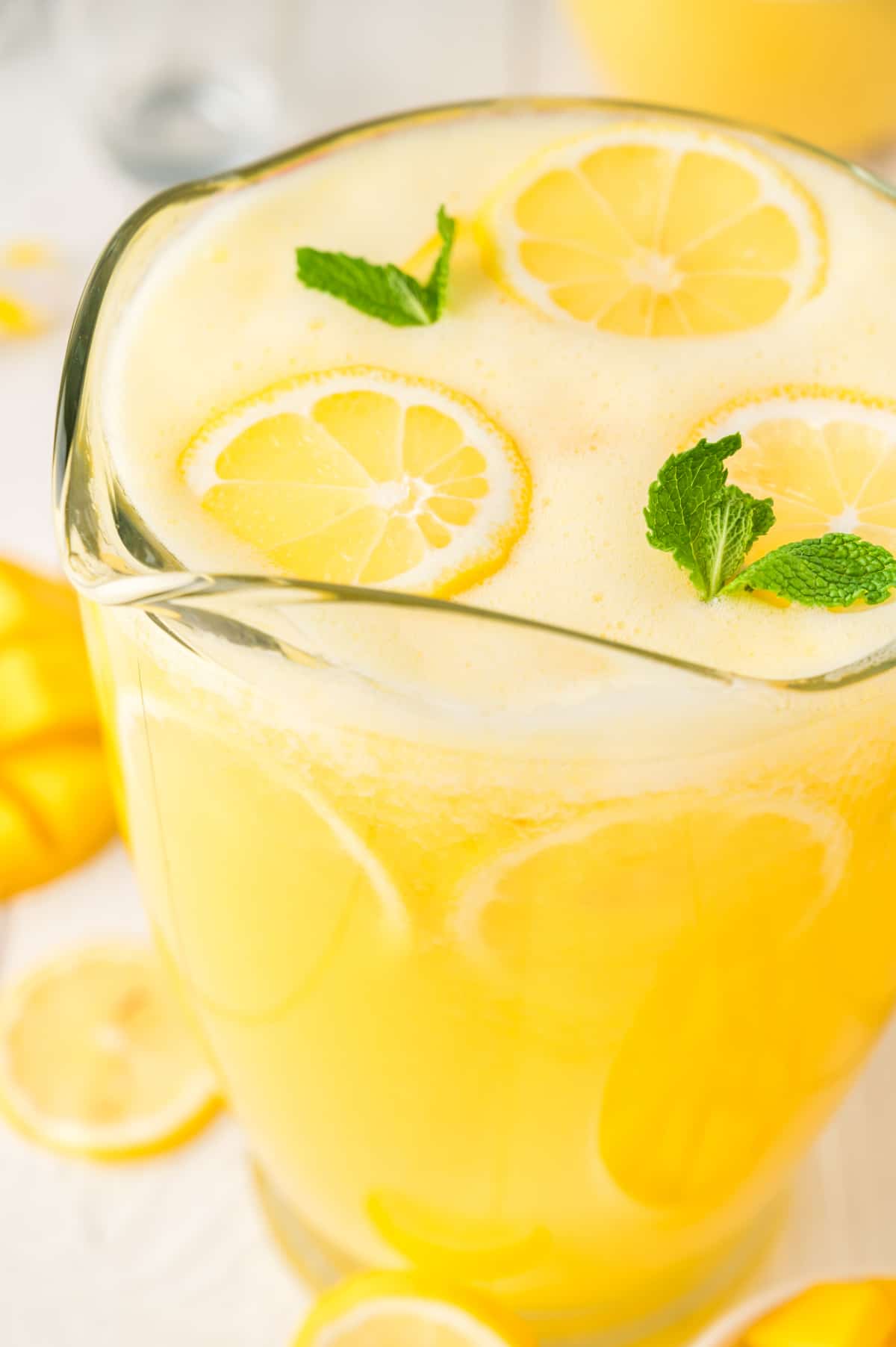 A clear pitcher filled with golden mango lemonade, garnished with lemon slices and fresh mint.