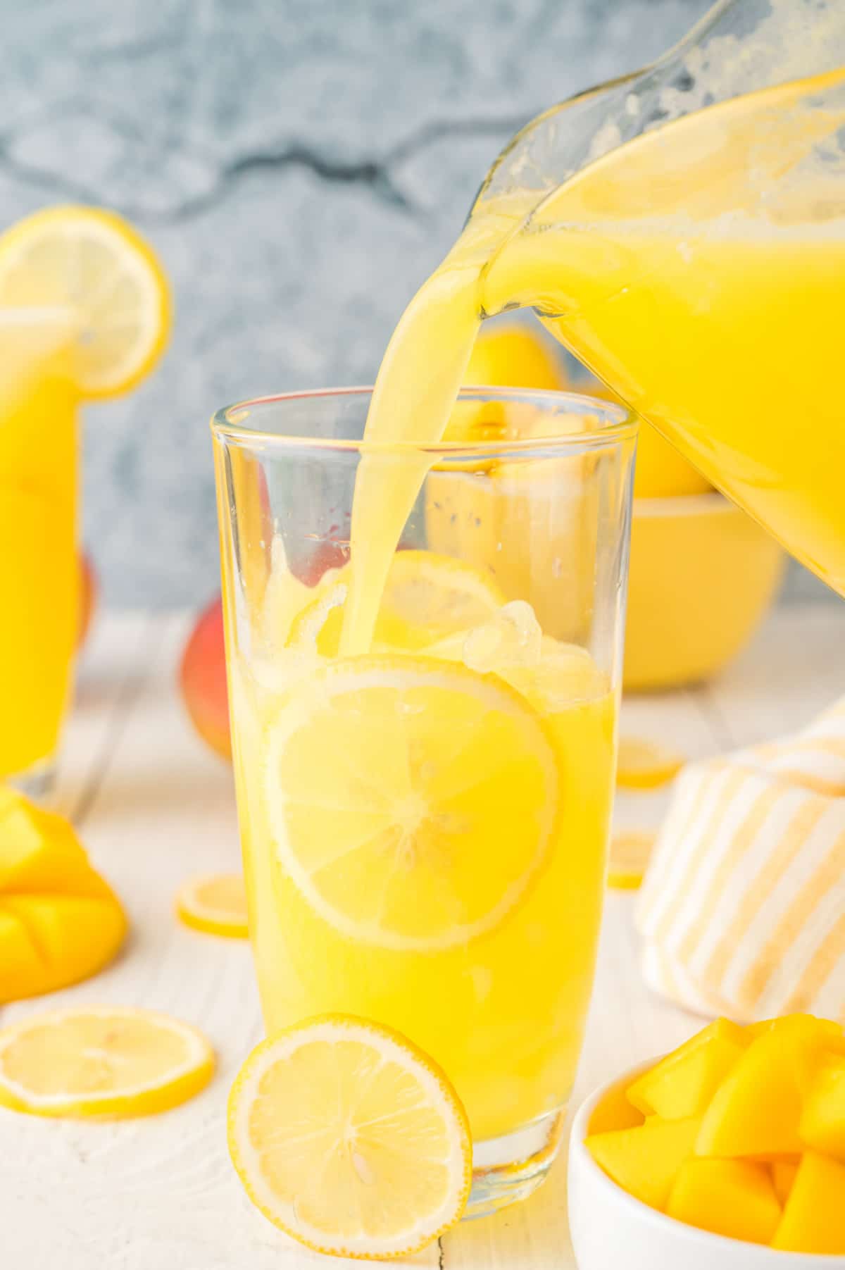 pouring mango lemonade from the pitcher into a tall glass filled with ice, with lemon and mango slices visible.