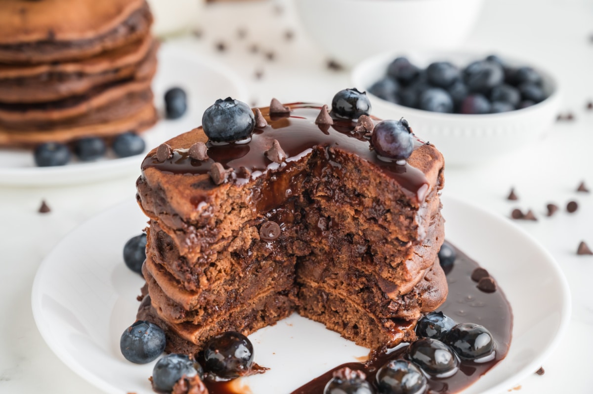 Close view of sliced double chocolate pancakes revealing melted chocolate chips inside, topped with ganache and fresh blueberries on a white plate. Cut stack of double chocolate pancakes showing melted chocolate chips inside with ganache and blueberries