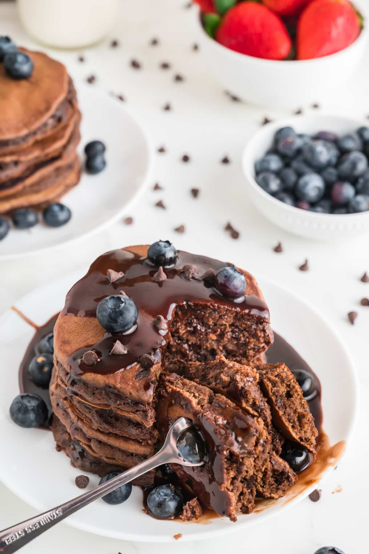 Stack of chocolate pancakes drizzled with chocolate ganache and fresh blueberries, partially sliced to reveal the moist chocolate center and melted chocolate chips inside. Chocolate pancakes topped with warm ganache and blueberries, sliced open to show soft chocolate interior