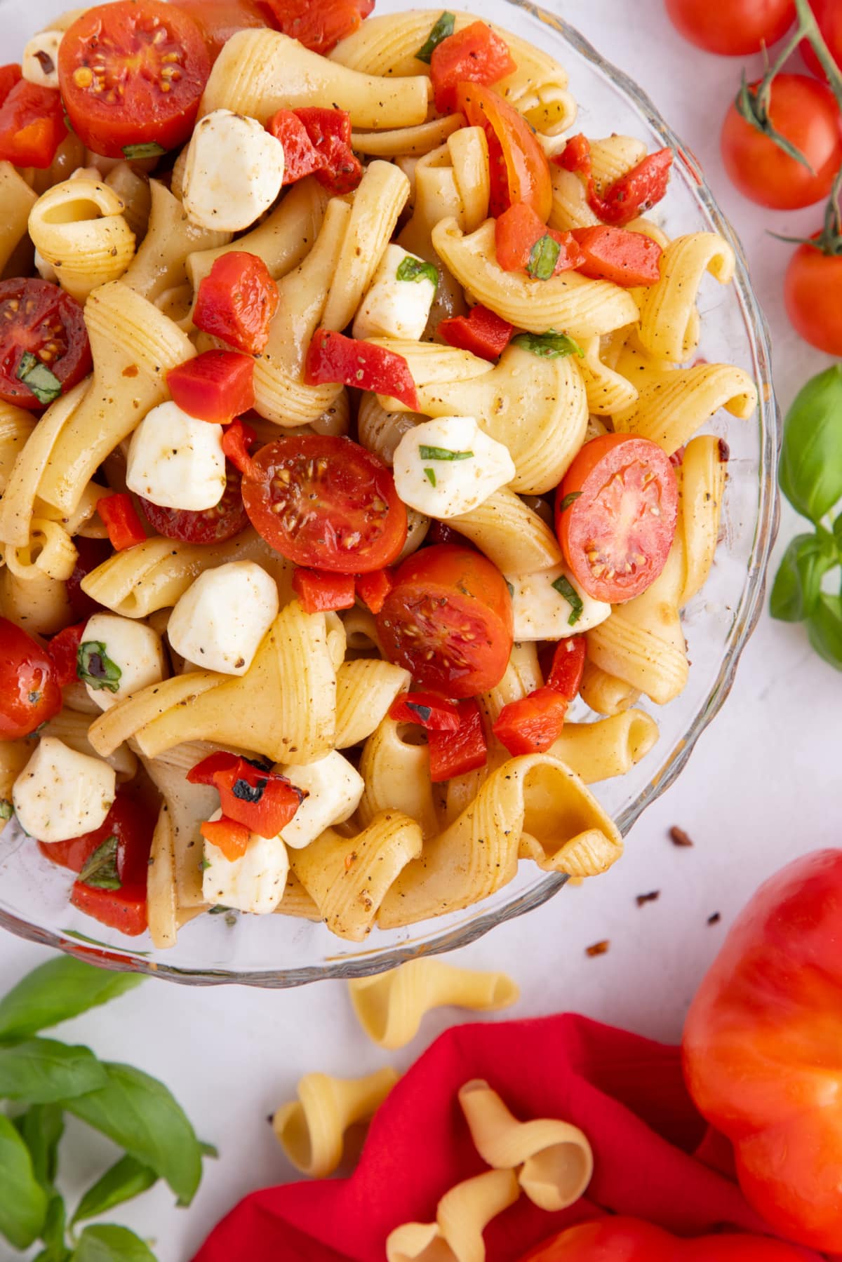 overhead view of Caprese Pasta Salad with Roasted Red Peppers Top-down view of caprese pasta salad with roasted red peppers in a clear glass bowl, showing the mix of pasta, mozzarella pearls, and basil.