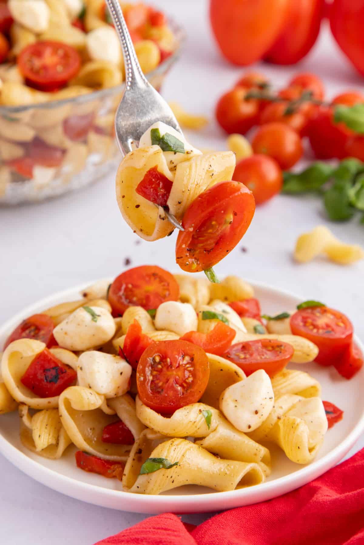 a bite of Caprese Pasta Salad with Roasted Red Peppers Close-up of a forkful of pasta salad lifted from the plate, showing a bite with mozzarella, pasta, tomato, and basil.
