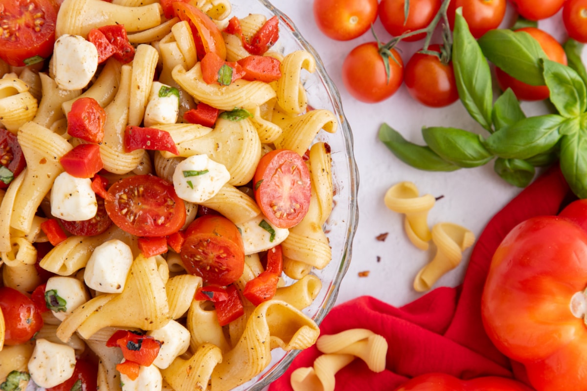 Caprese Pasta Salad with Roasted Red Peppers with tomatoes and fresh basil Overhead shot of the finished pasta salad in a serving bowl, styled with fresh cherry tomatoes and basil leaves on the side for garnish.