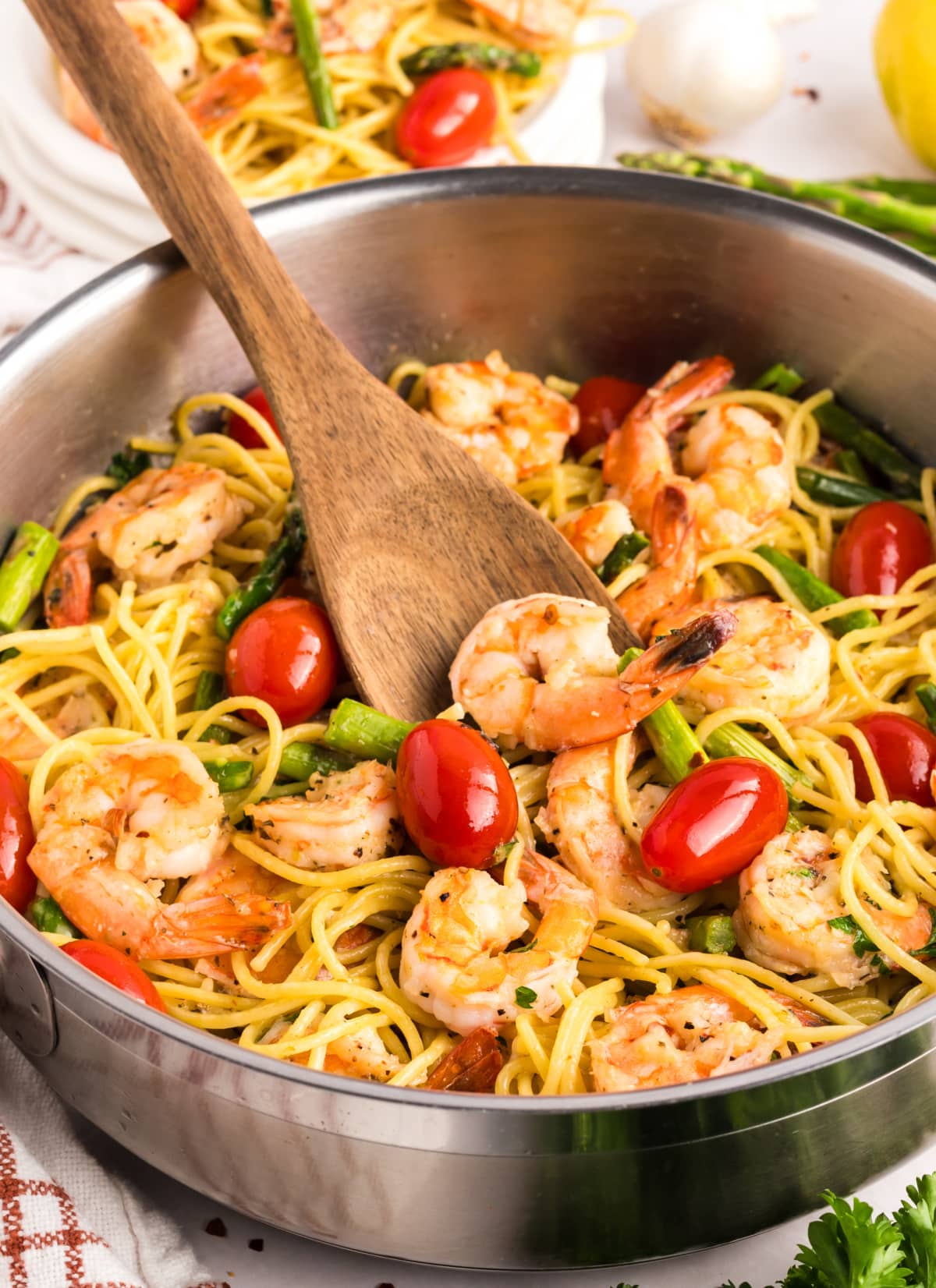 stirring the Garlic Shrimp Pasta with Asparagus and Tomatoes Wooden spoon stirring garlic shrimp pasta in a skillet, with shrimp and vegetables visible throughout.