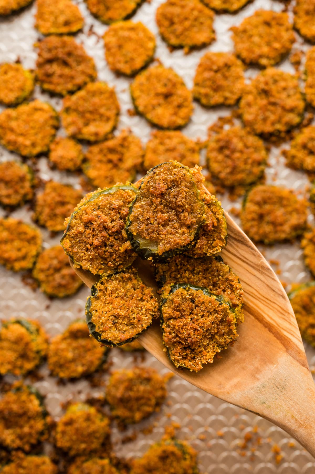 Close-up of oven fried dill pickles on a wooden spoon with a full baking tray in the background.