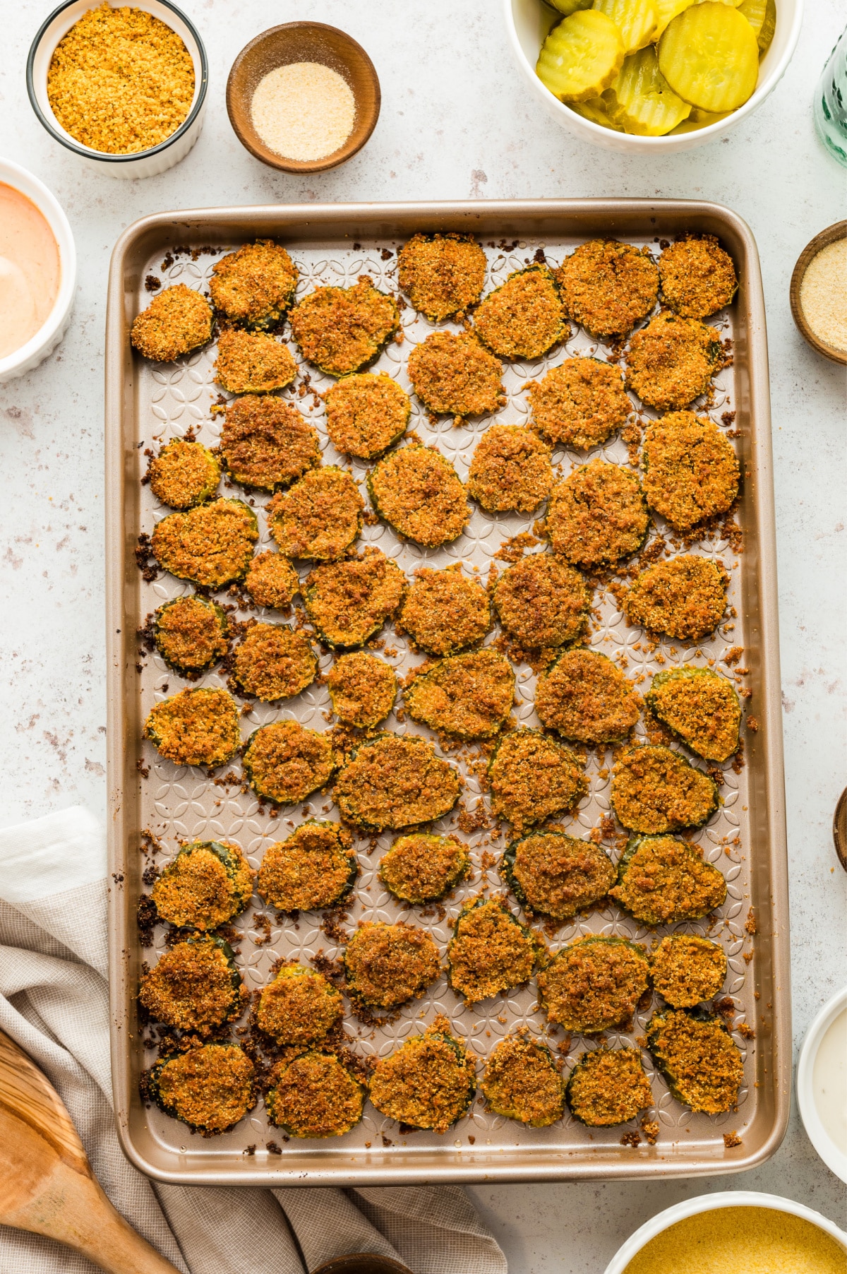 Baked oven fried dill pickles on a tray, surrounded by key ingredients used in the recipe.
