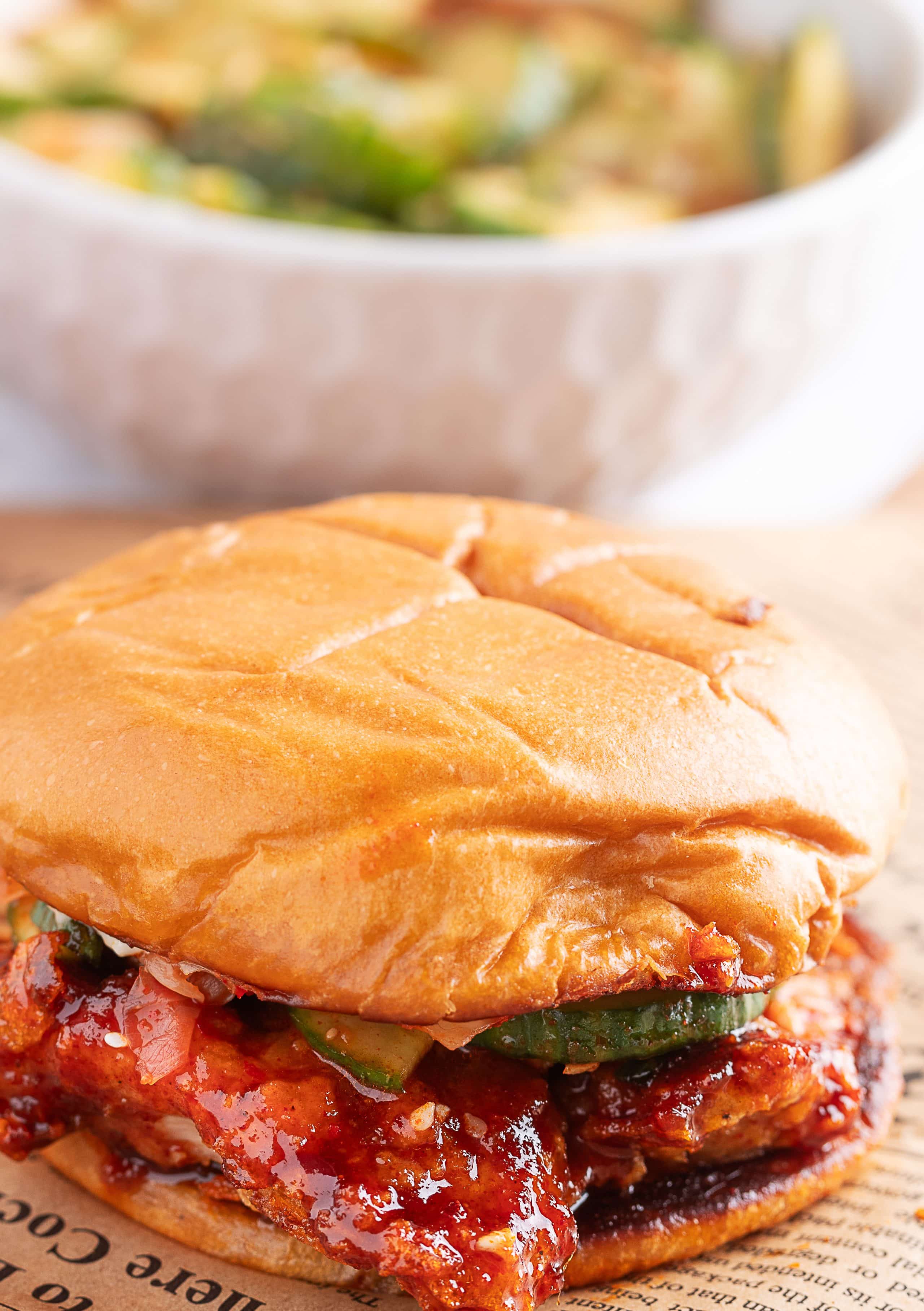Casual shot of finished sandwich on kitchen counter with cucumber salad Gochujang Chicken Sandwich sitting on a counter, ready to serve with cumcumber salad in the background