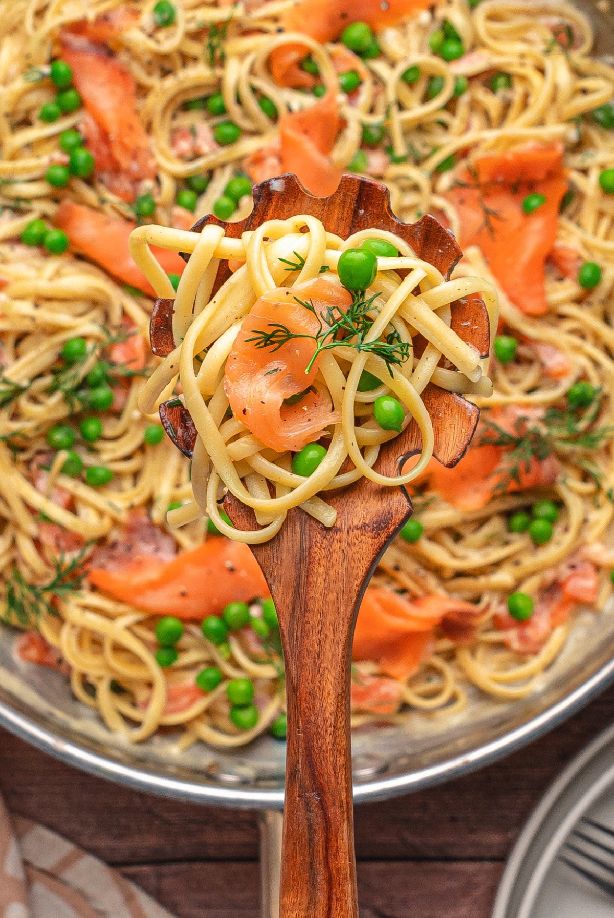 Smoked salmon pasta with lemon and dill being served from a skillet. Wooden spoon lifting creamy smoked salmon pasta from a skillet, top view.