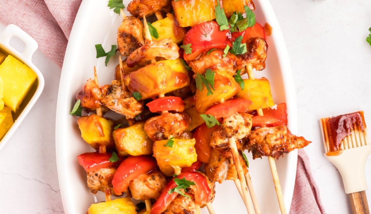 Baked BBQ chicken skewers on a platter with fresh pineapple and a BBQ sauce brush