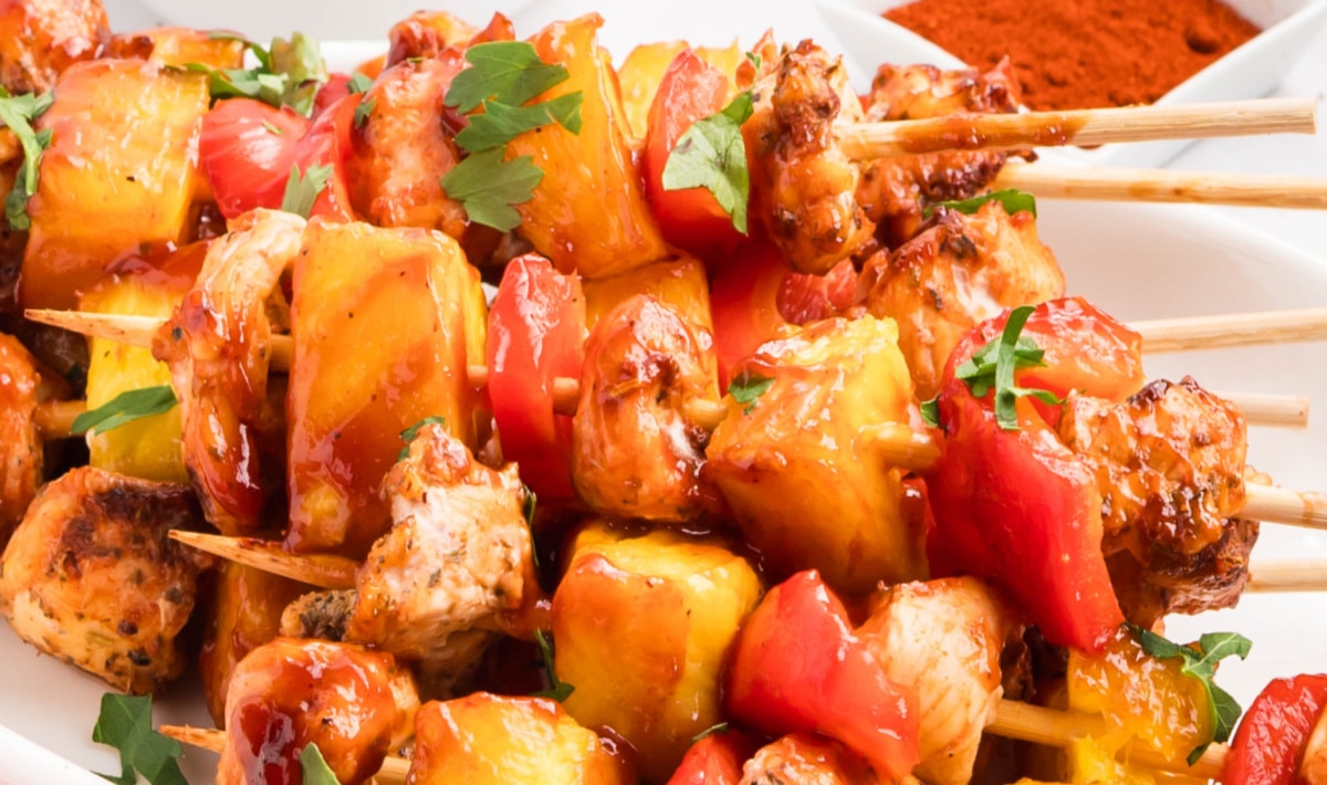 Side view of BBQ chicken skewers arranged on a white platter with pineapple and peppers
