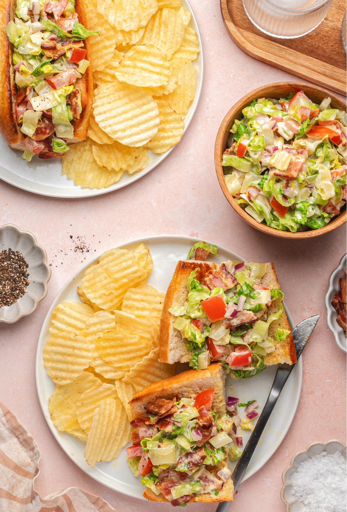 Flat lay of chopped BLT hoagie sandwiches on plates, cut in half with potato chips, water, and ingredients scattered around