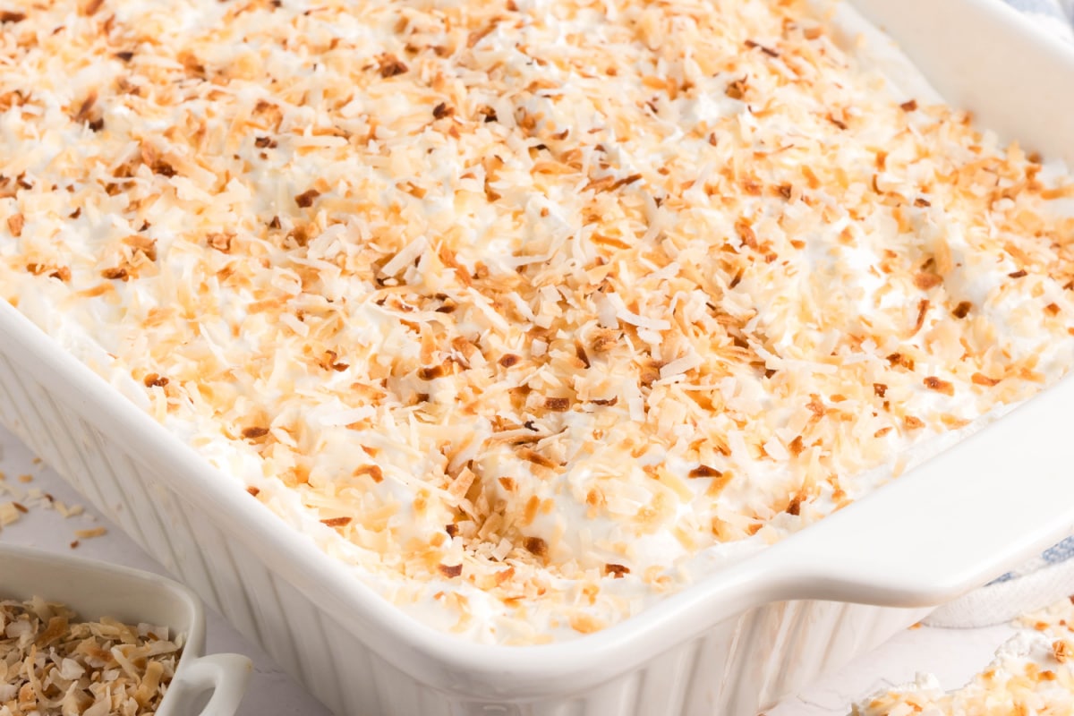 Side view of the full coconut poke cake topped with whipped cream and toasted coconut in a glass baking dish.