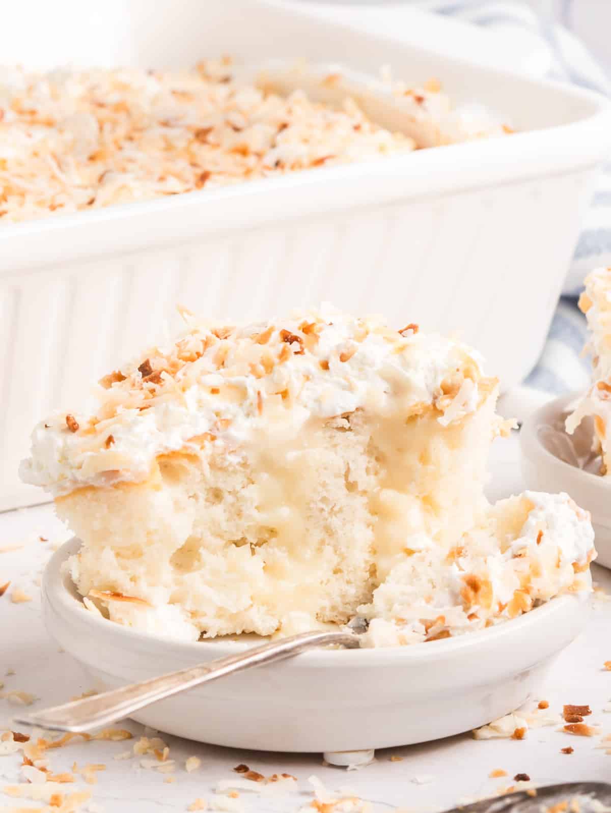 A slice of coconut poke cake on a plate with a spoonful lifted, full cake in the background.