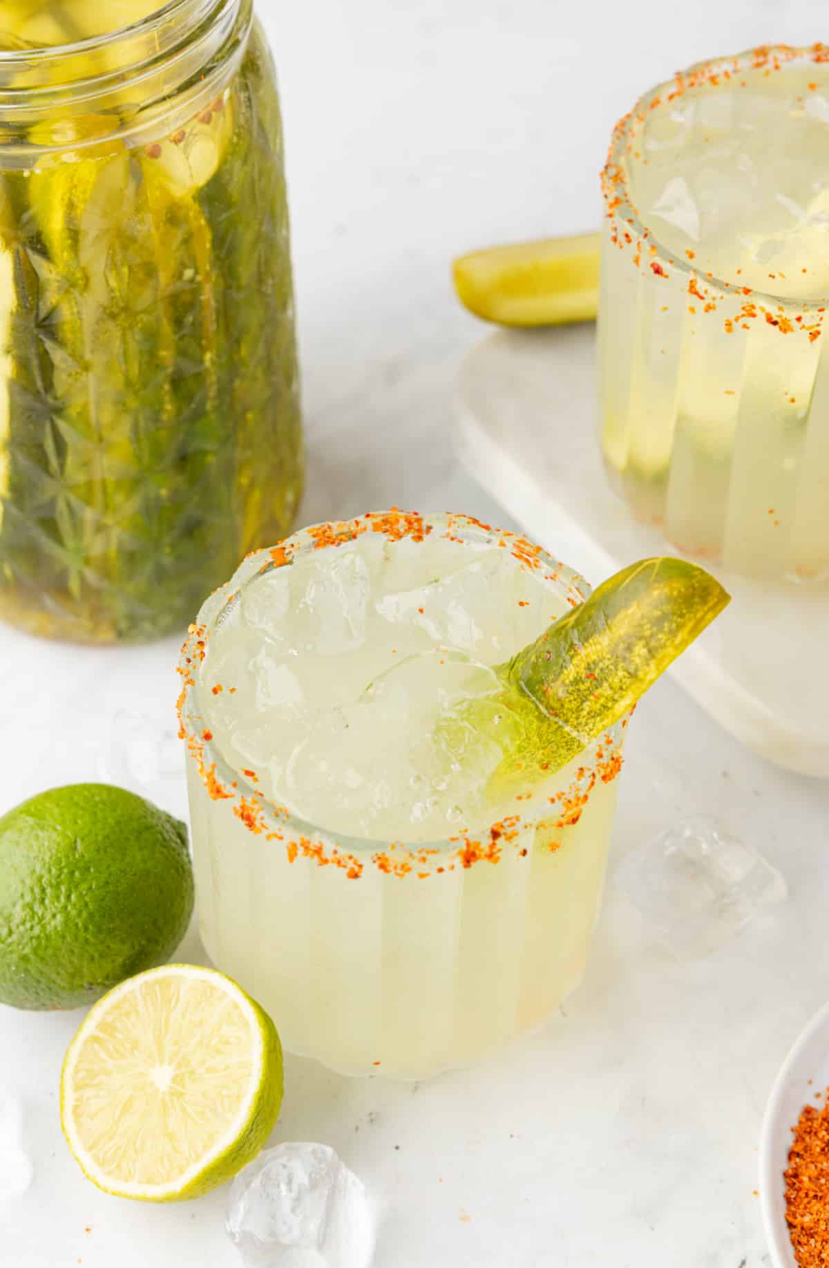 Two Pickle Margaritas with Garnishes and Ice Two dill pickle margaritas with a pickle jar, lime, and ice on the table