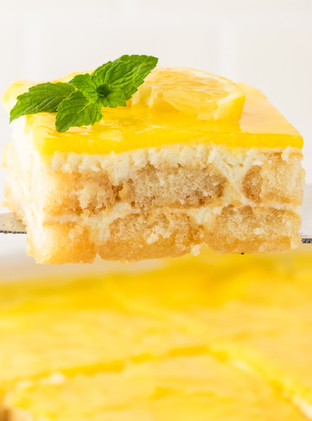 A close-up of a lemon tiramisu slice held on a spatula above the cake dish.
