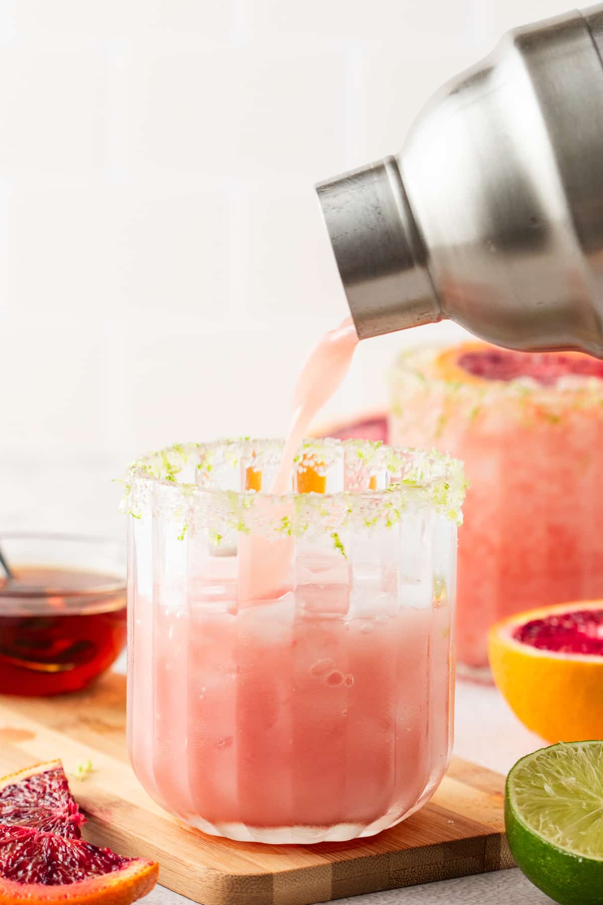 Pouring a Tropical Blood Orange Margarita Pouring a coconut blood orange margarita into a glass with lime zest salt rim and blood orange slices nearby.